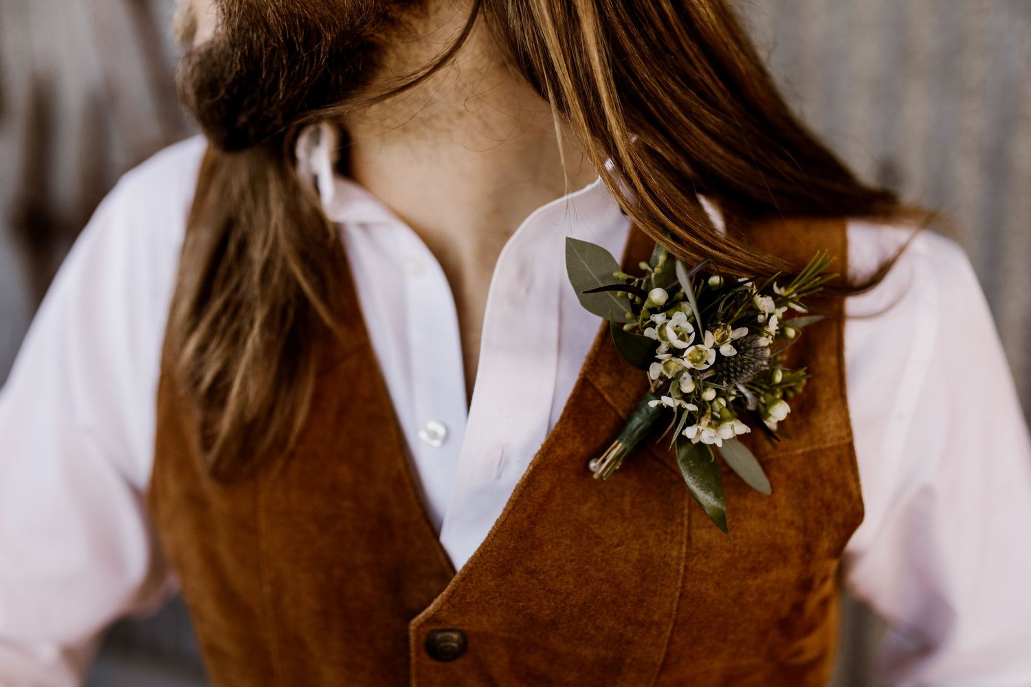 Close-up of rustic greenery boutonniere pinned to groom wearing brown suede vest and white shirt.