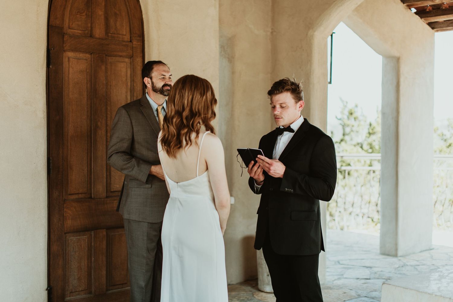 Intimate elopement ceremony with bride and groom listening to officiant read vows at rustic venue.