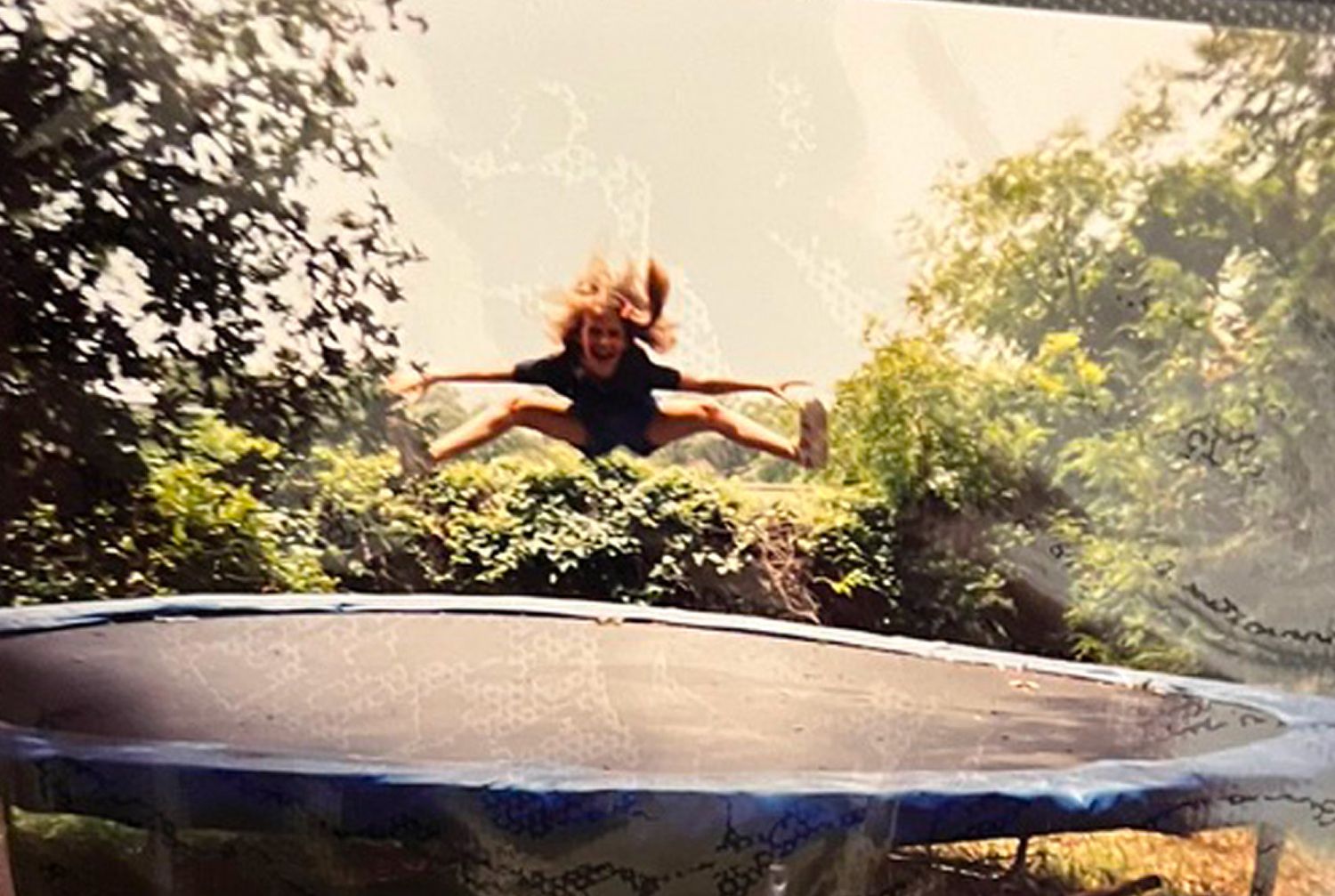 A young girl jumps joyfully on a trampoline surrounded by lush green trees in a backyard.