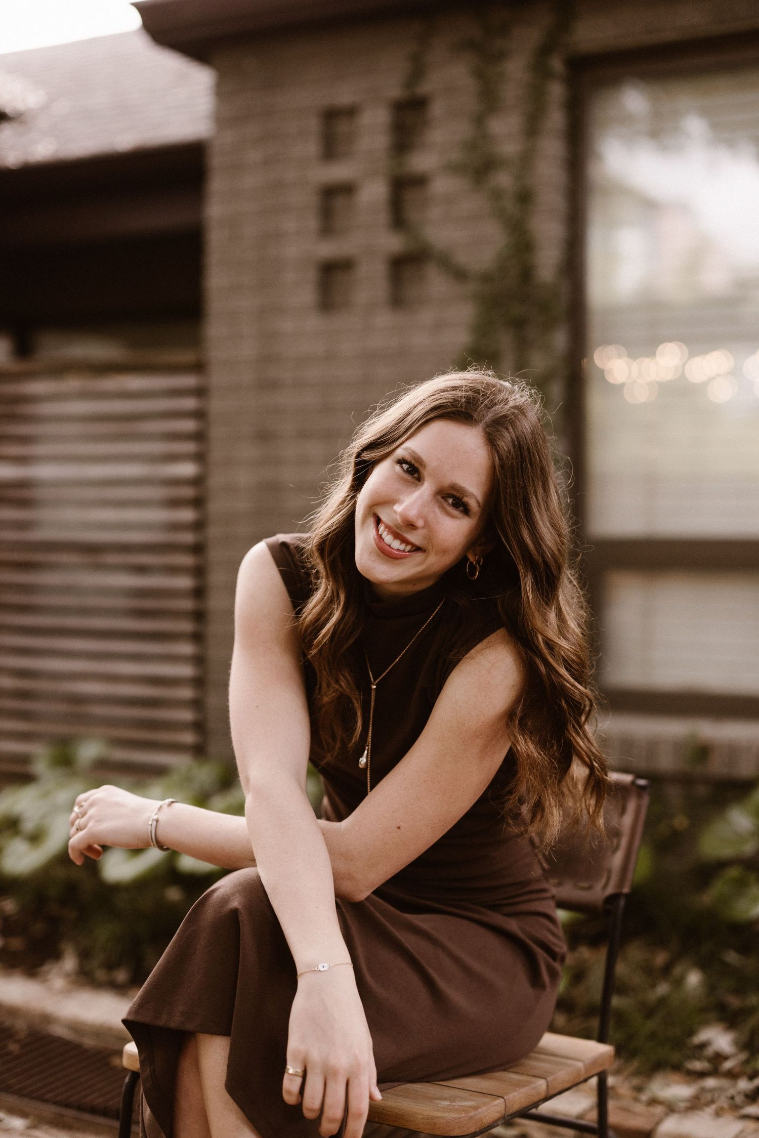 Smiling young woman in a brown sleeveless dress sitting on a bench outdoors with bokeh lights behind her.