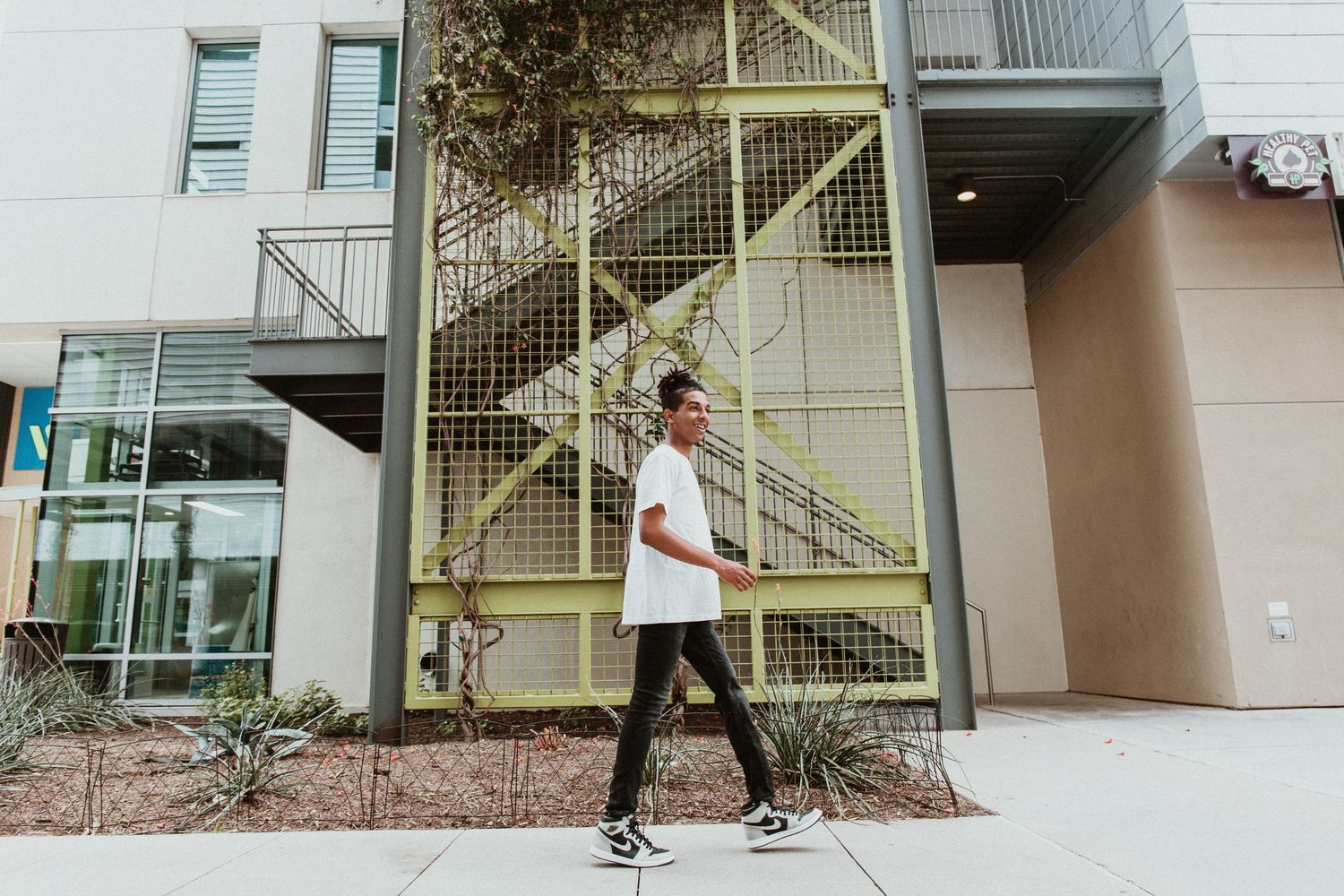 A young man in white shirt and black pants walks past a modern building with a yellow fire escape staircase.