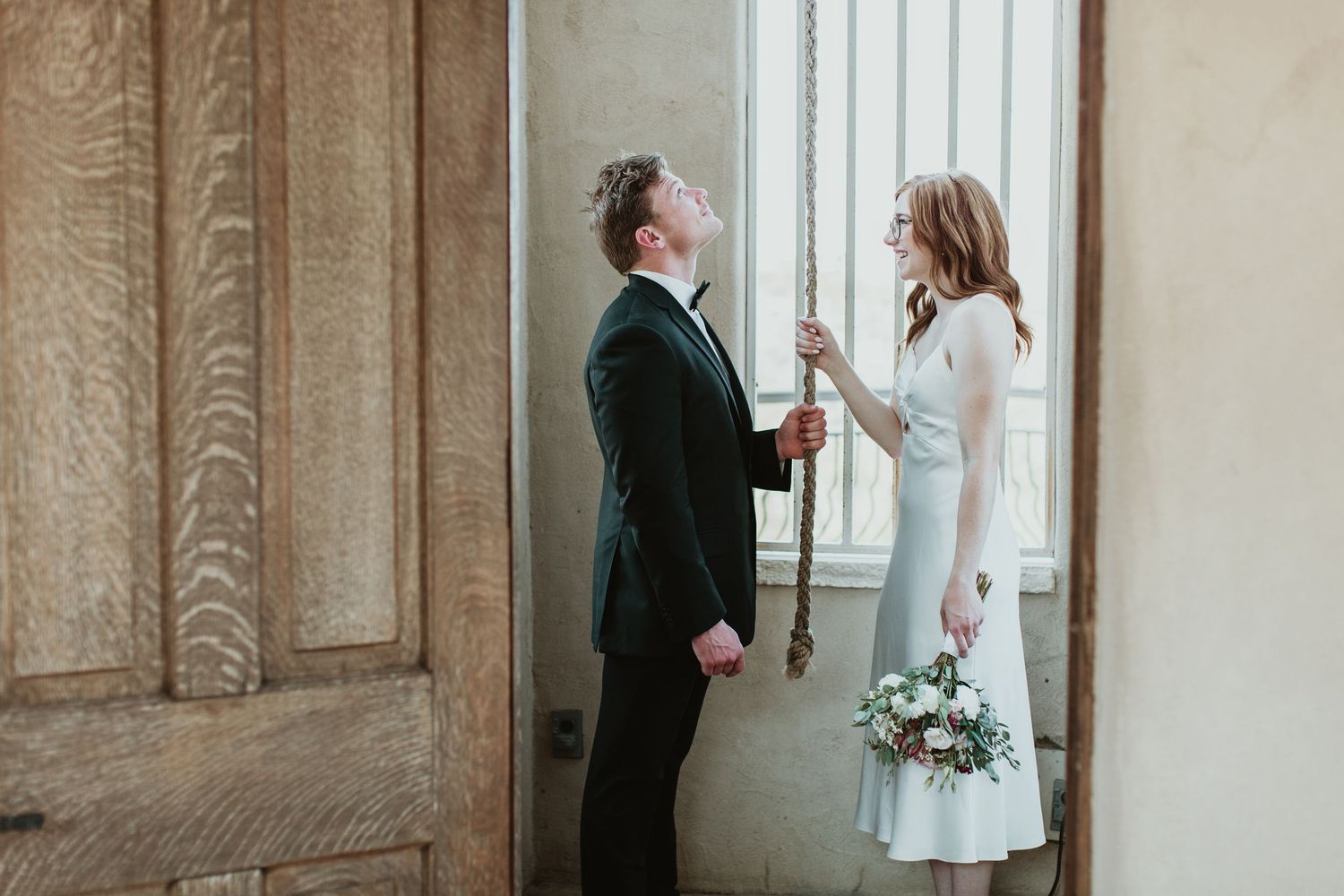 Bride and groom stand by a vintage window, she holds a bouquet, as he pulls a decorative rope in an elegant venue.