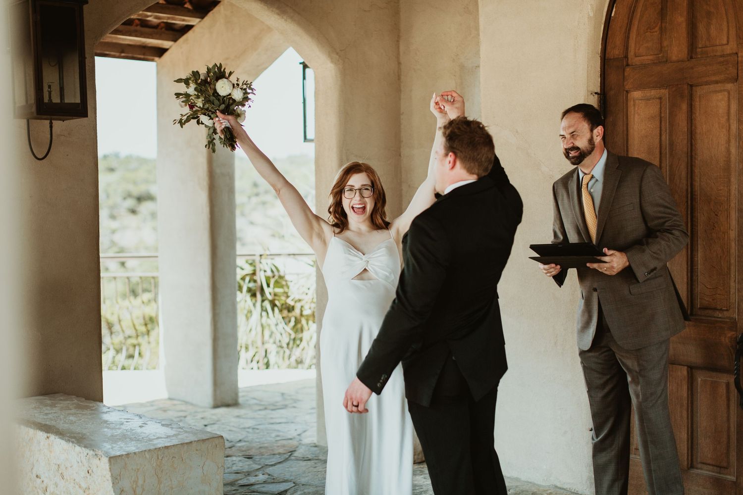 Joyful bride raises bouquet during intimate outdoor wedding ceremony with groom and officiant at rustic venue.