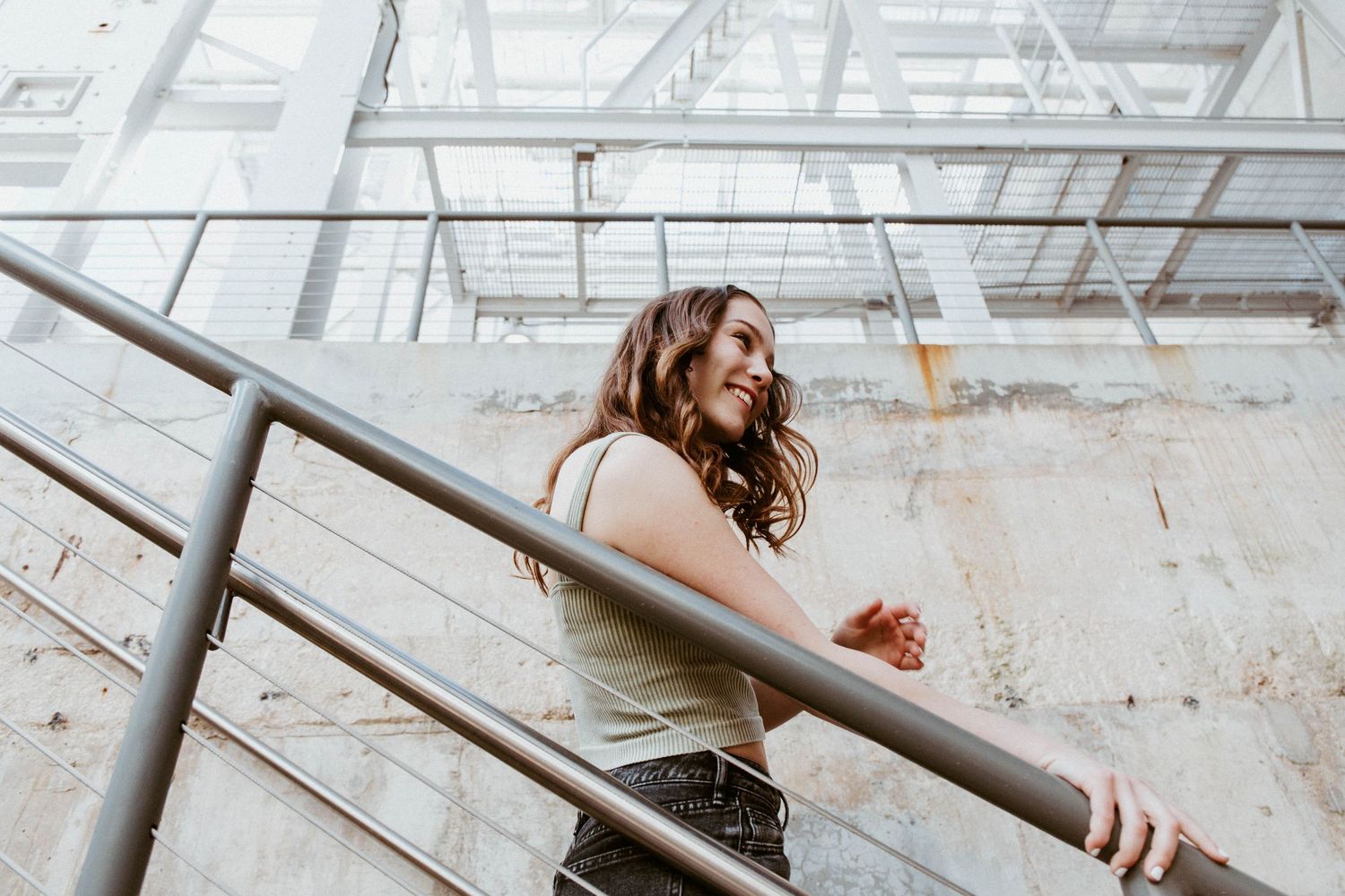 Young woman with wavy brown hair leaning on a metal railing inside an industrial building with glass ceiling.