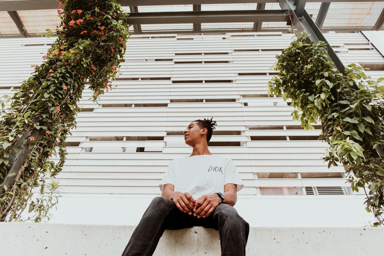 Young man in white t-shirt sitting outdoors near hanging green plants against a white slatted wall.