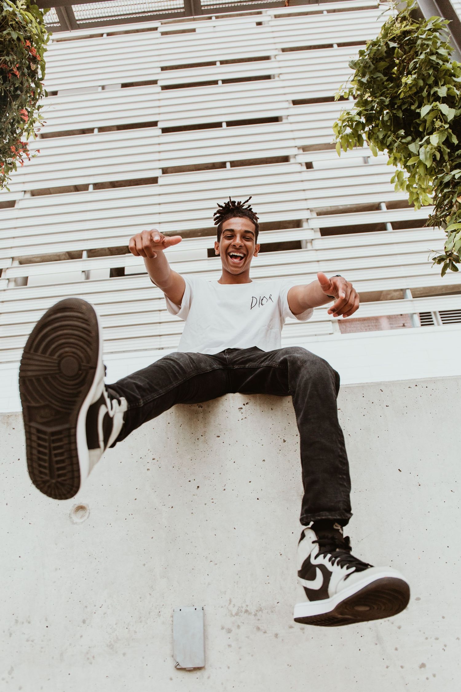 Young man sitting on a wall laughing, wearing white tee, black jeans, and Jordan sneakers, kicking one leg up.