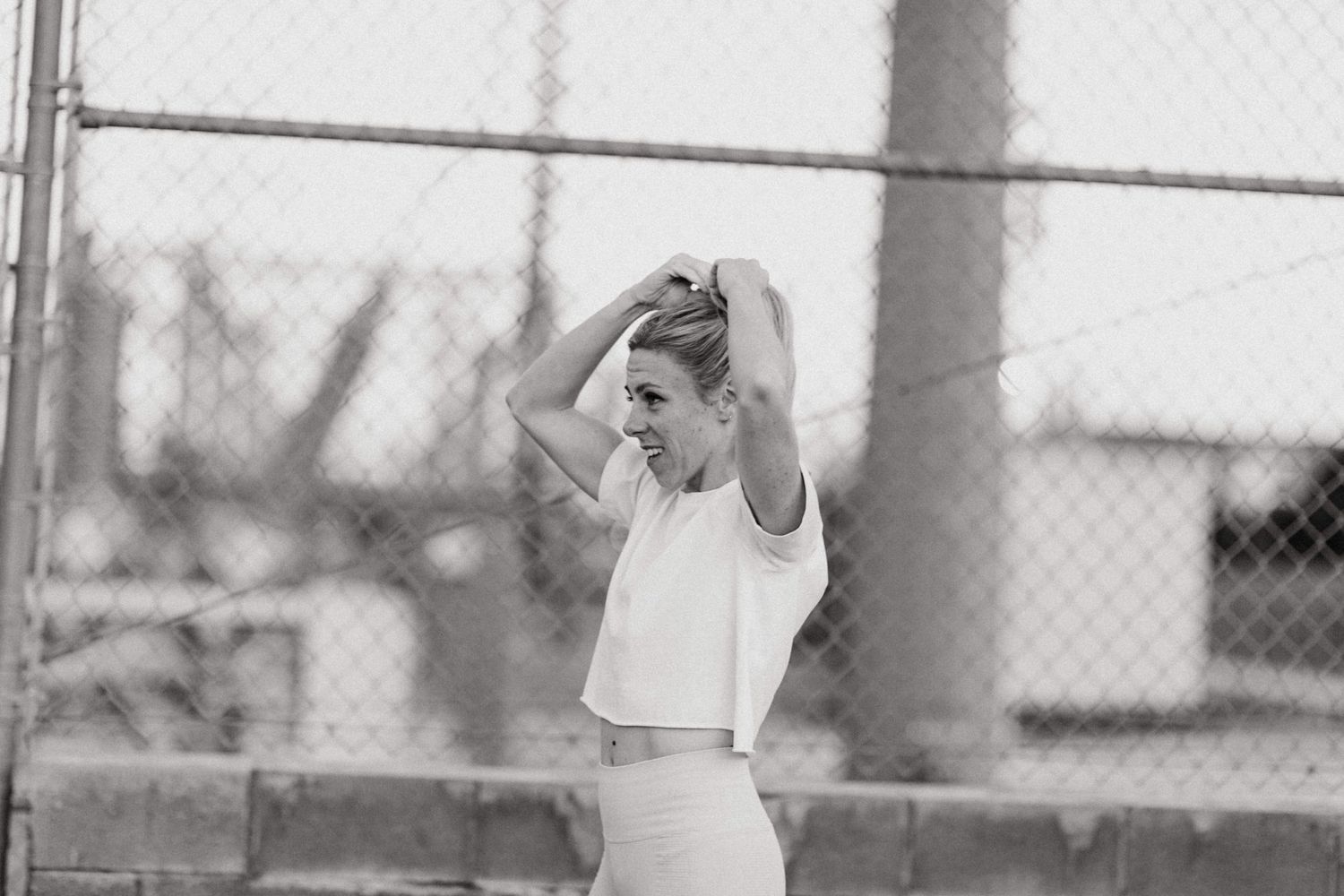 Black and white photo of a young woman in a white crop top adjusting her hair near a chain-link fence outdoors.
