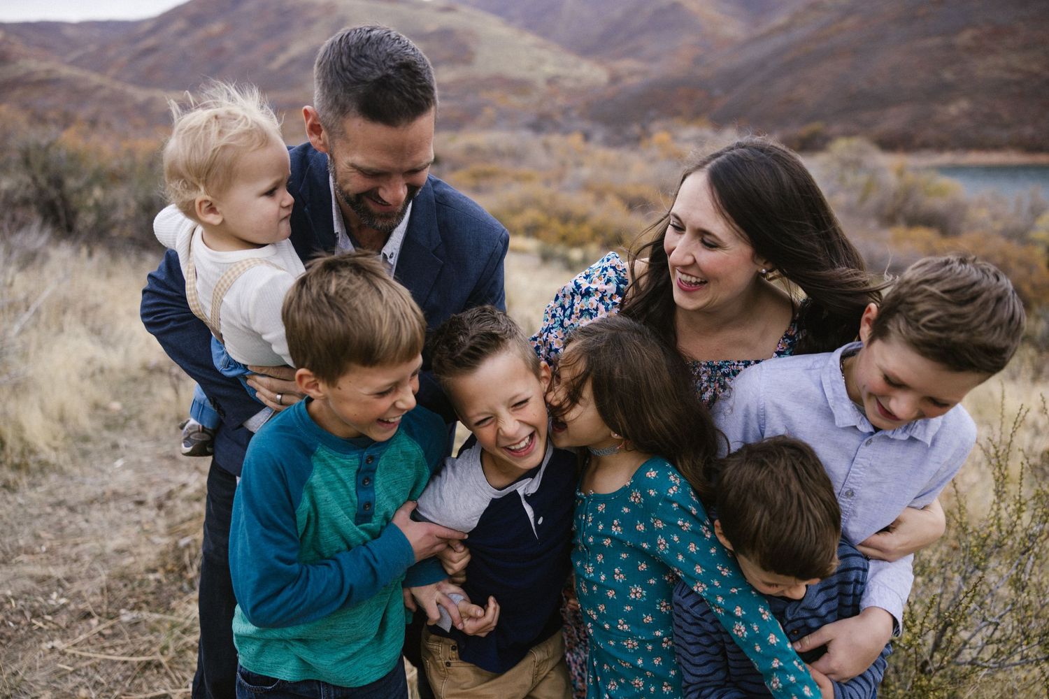 Happy family with five children laughing and embracing outdoors near a scenic lake and hills.