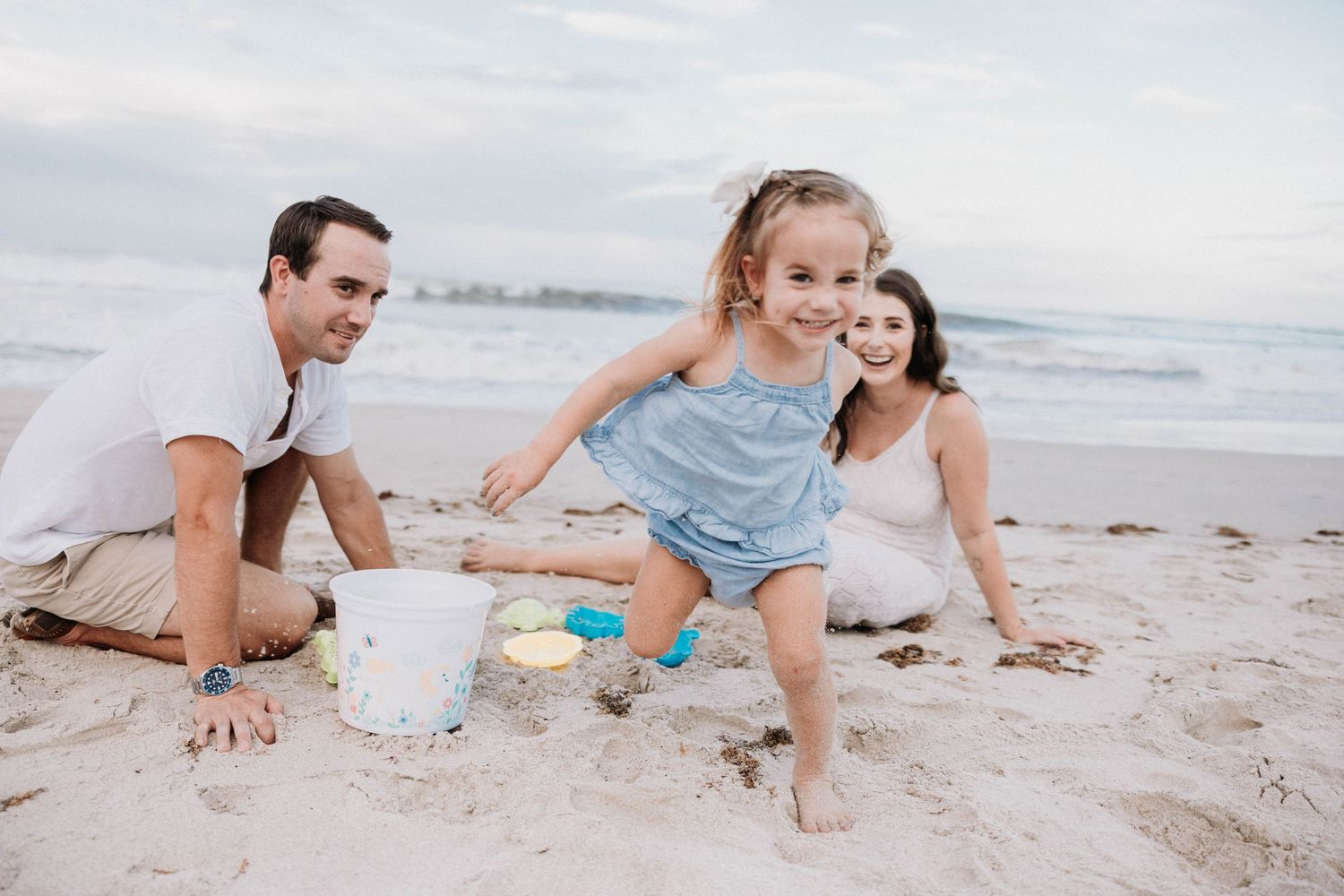 Young family with toddler girl playing in sand on a beach with colorful toys.