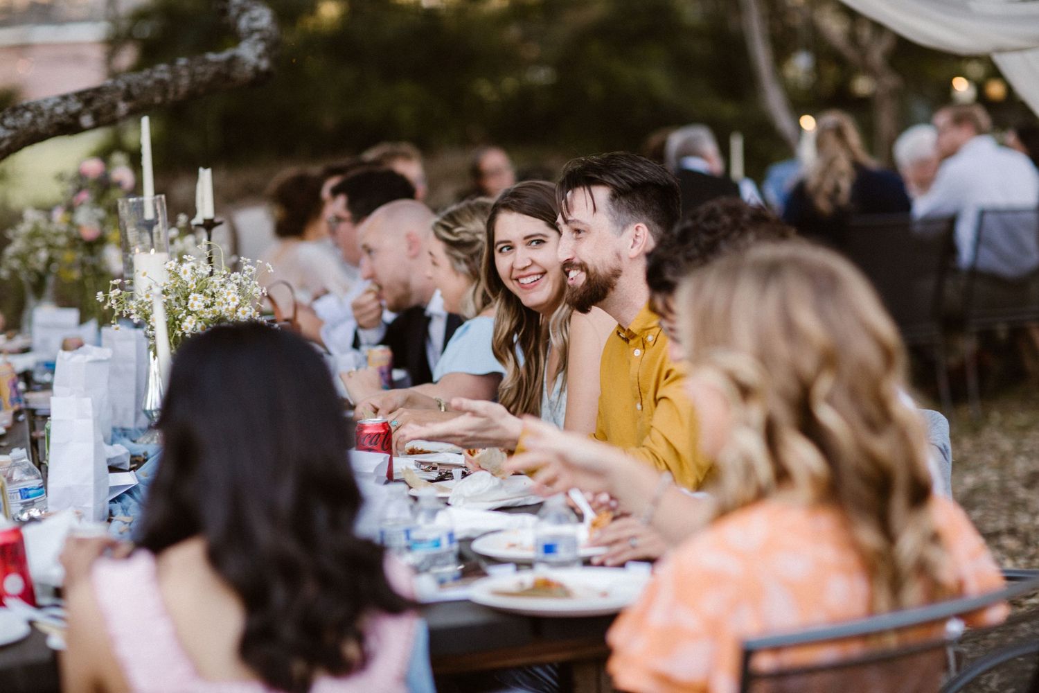 Guests laugh and enjoy dinner together at long tables during an outdoor wedding reception.