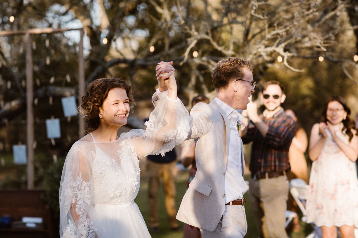 Smiling bride and groom dancing together at outdoor wedding reception under string lights with guests.