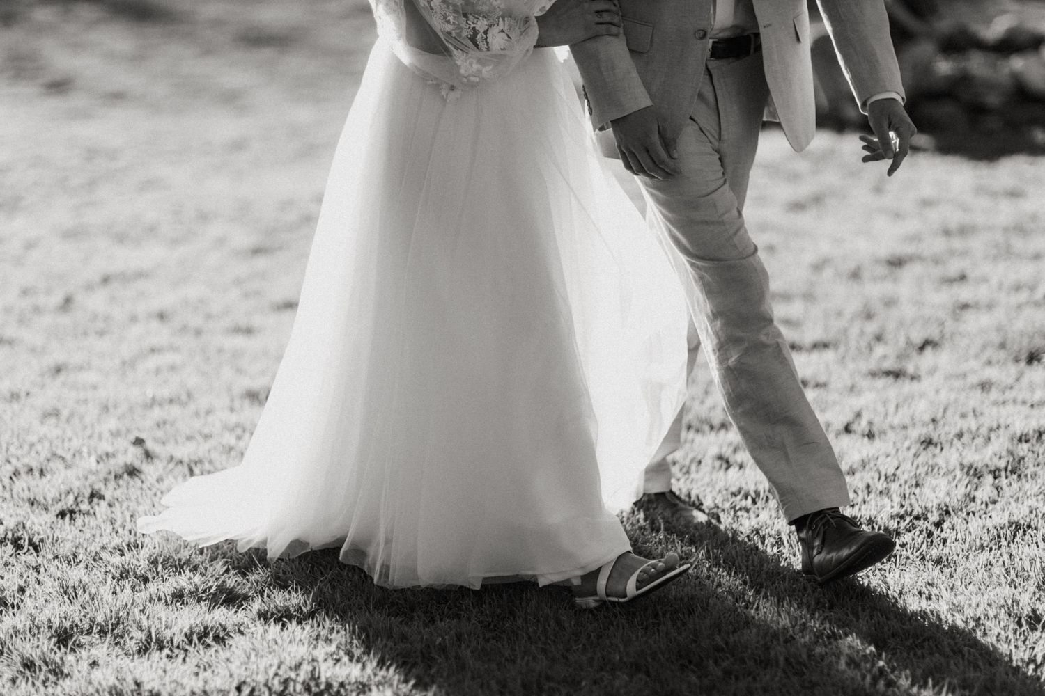 Black and white photo of bride in flowing tulle gown and groom walking on grass at wedding.