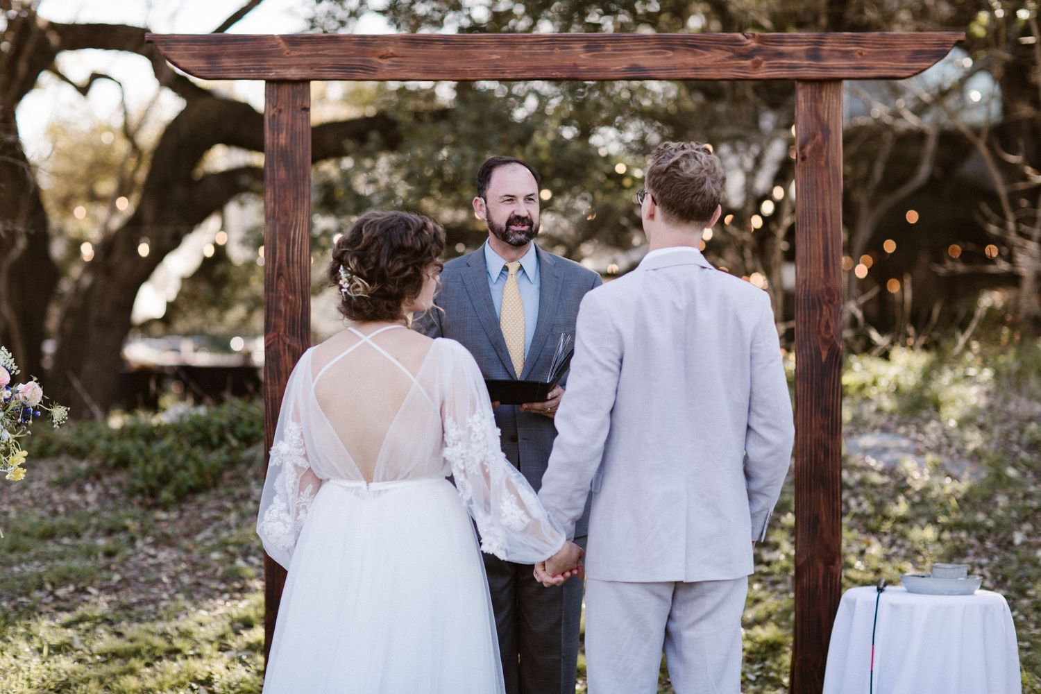 Bride and groom hold hands before officiant under rustic wooden arch at outdoor garden wedding ceremony.