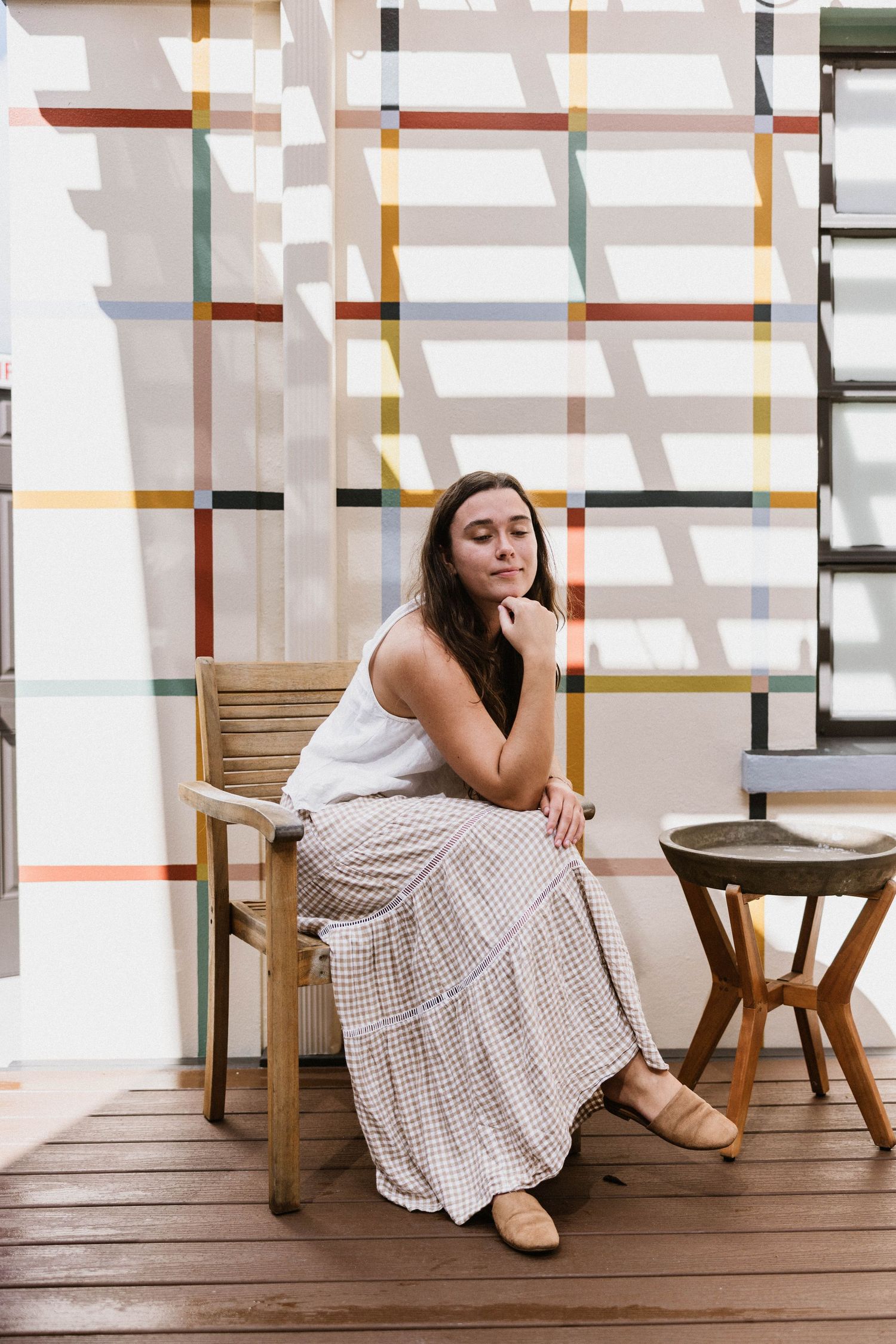 A young woman in a flowy skirt sits thoughtfully on a wooden chair beside a colorful geometric wall.