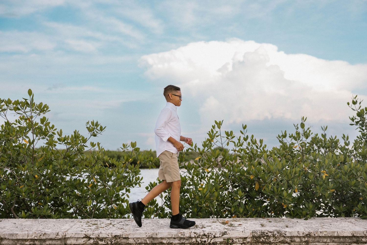 Boy in white shirt and khaki shorts walks along a stone wall with cloudy sky behind him.