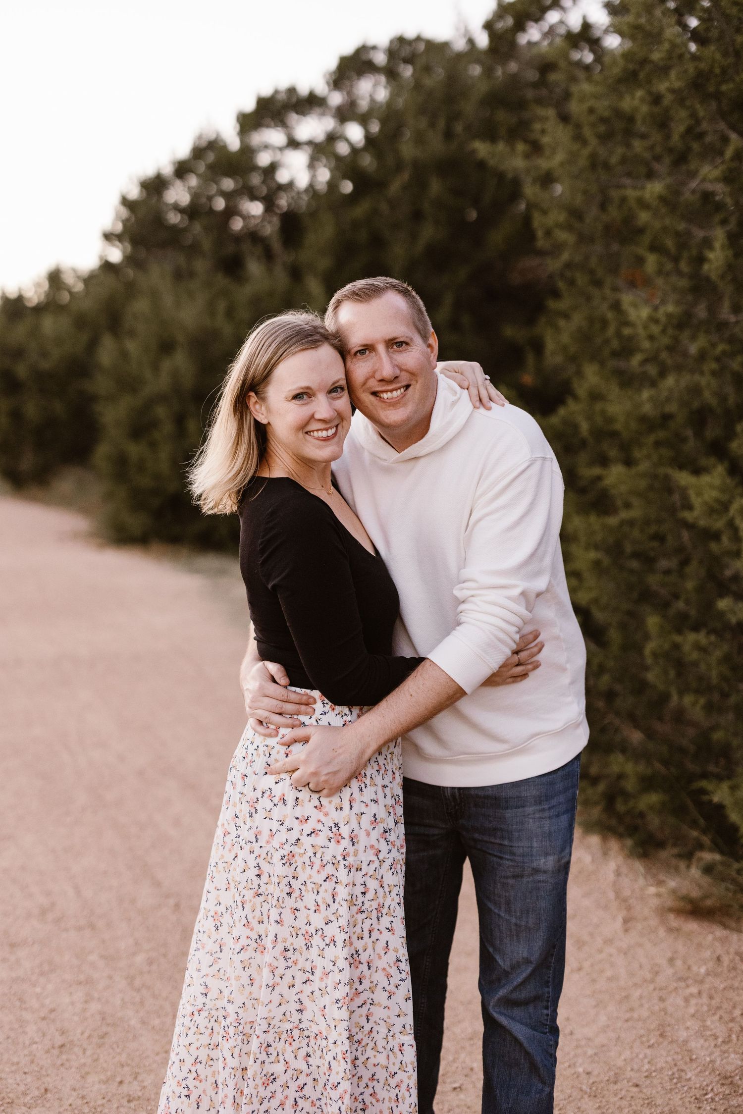 Smiling couple embracing on a dirt path surrounded by green trees during an outdoor photo session.