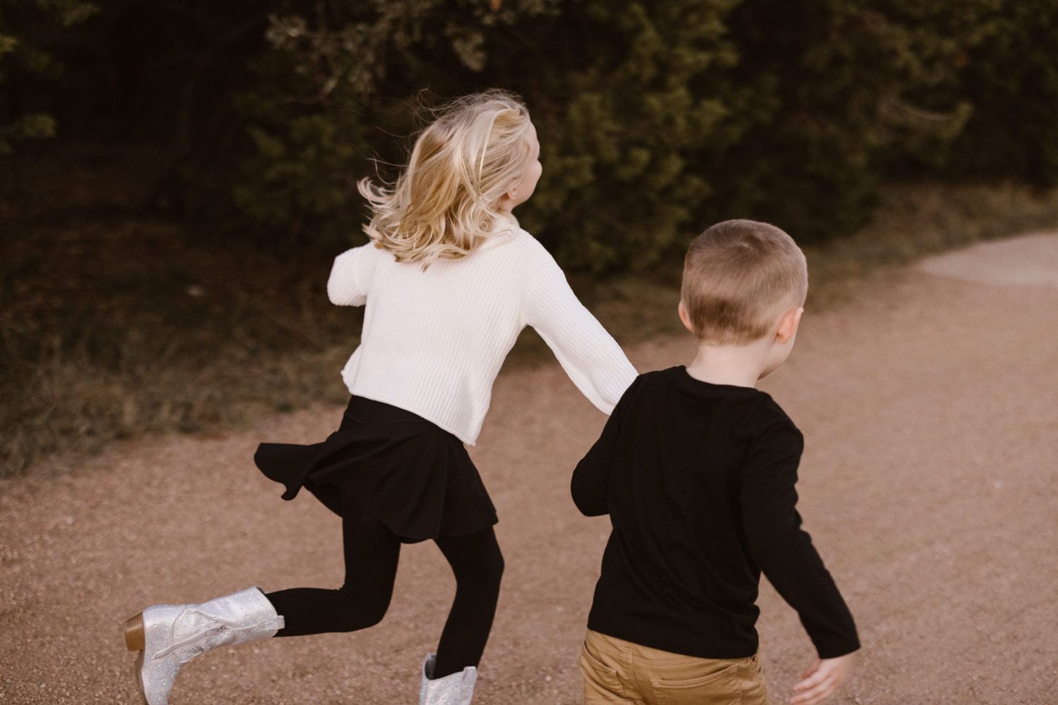Two young children running together down a dirt path near green trees.
