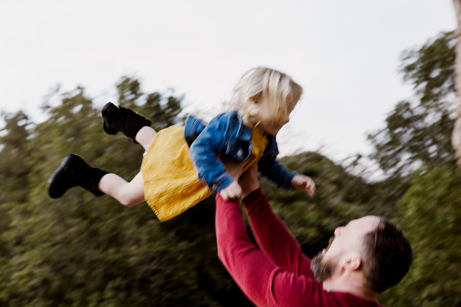 Father joyfully lifting his young child high into the air outdoors among trees.