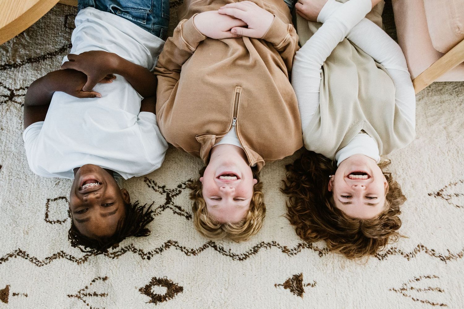 Three smiling teens lying on their backs on a patterned rug indoors.