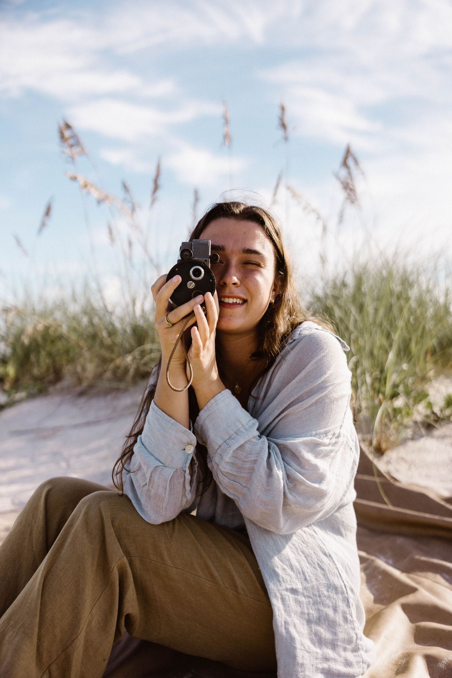 Smiling woman holding vintage film camera on sandy beach with sea oats behind her.