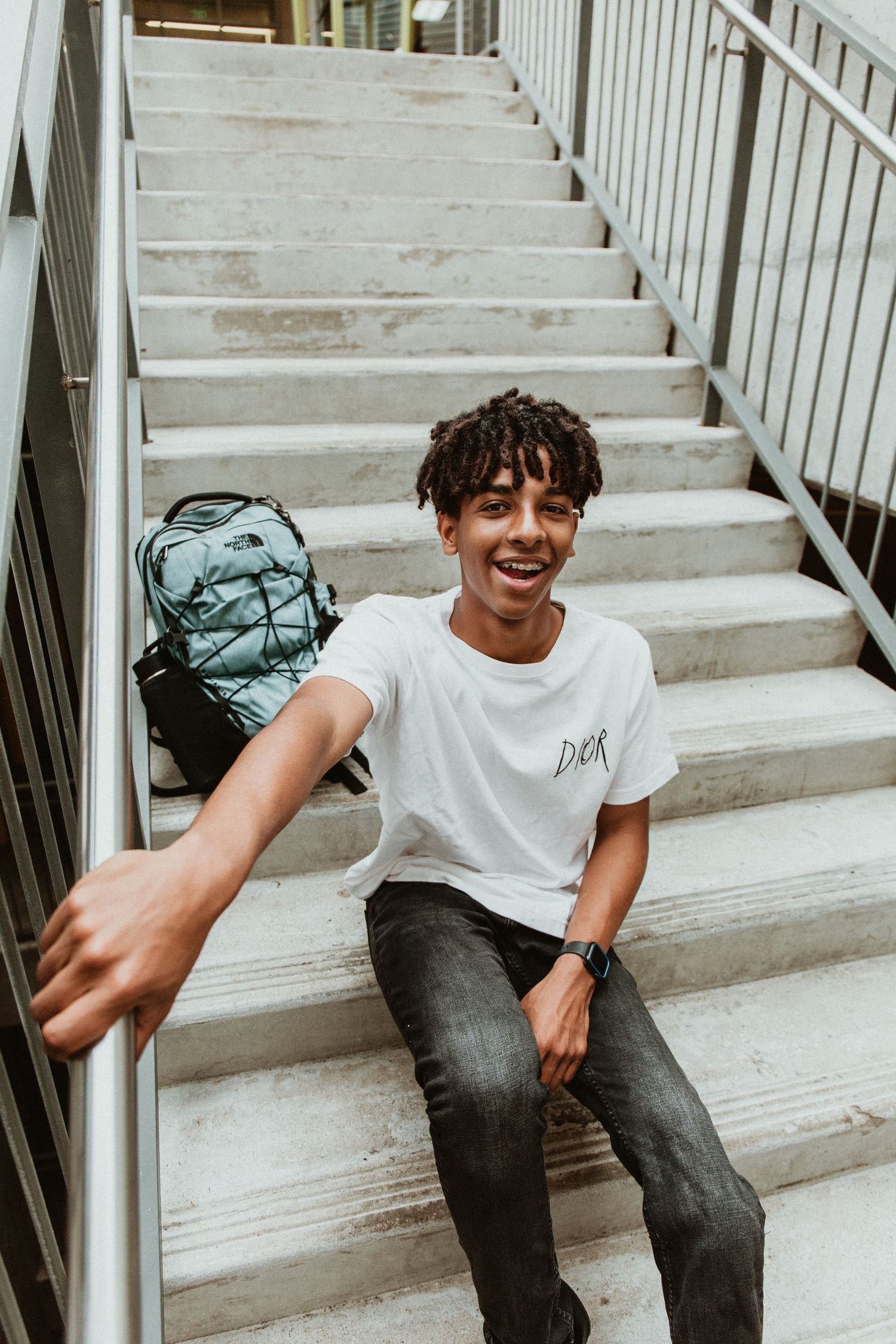 Smiling young man in white Dior tee sitting on stairs with backpack, reaching toward camera.