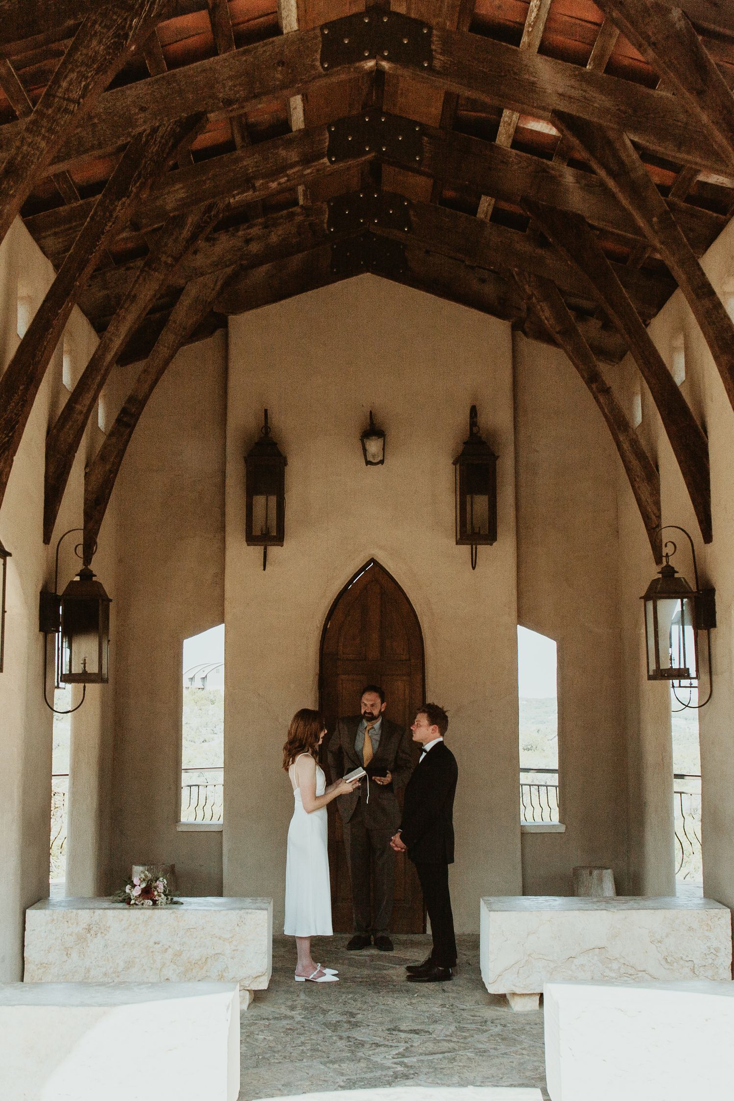 Couple exchanges vows under wooden arched ceiling in elegant chapel with gothic door and lantern sconces.