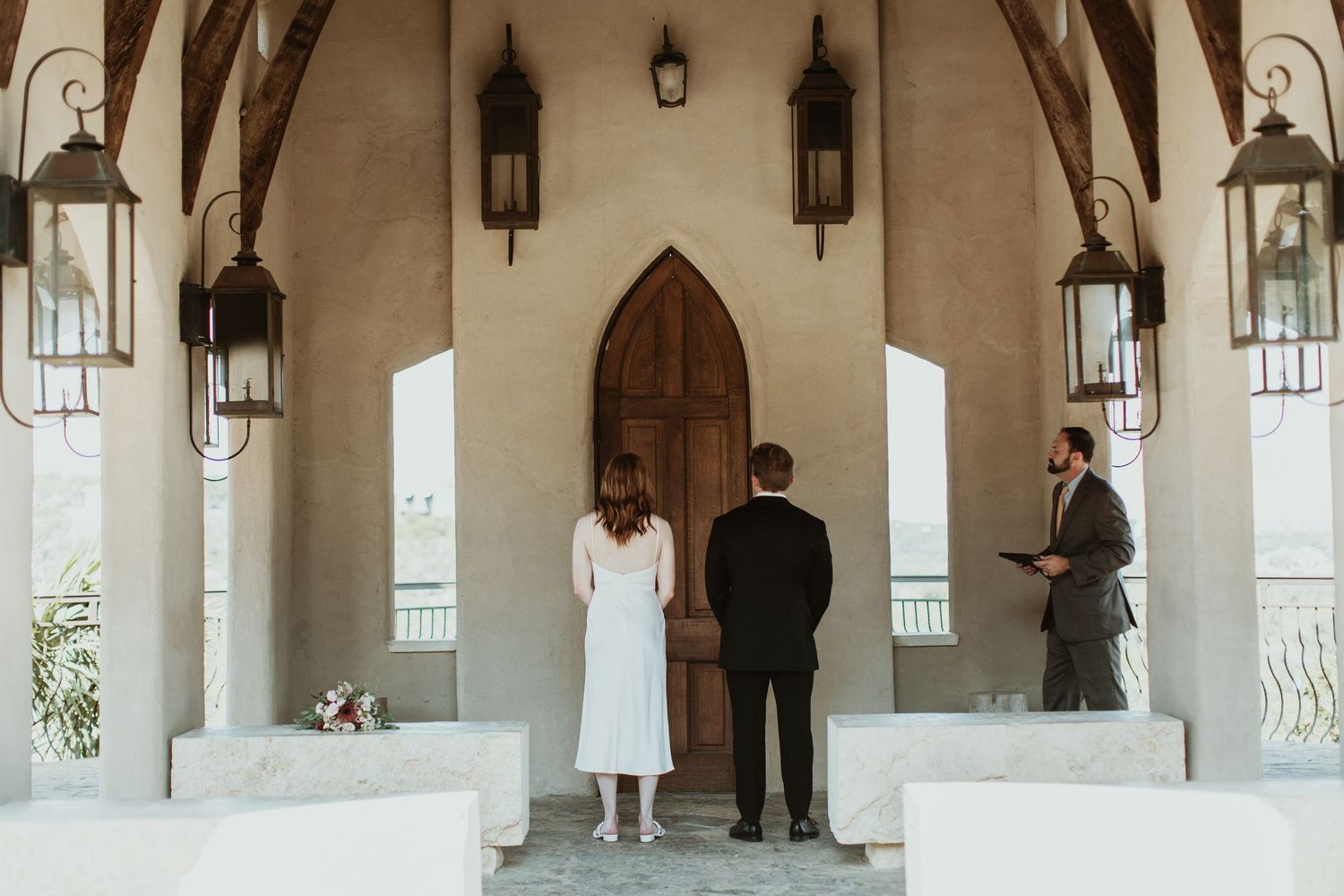 Couple stands before ornate chapel doors with lanterns during intimate outdoor wedding ceremony with officiant.