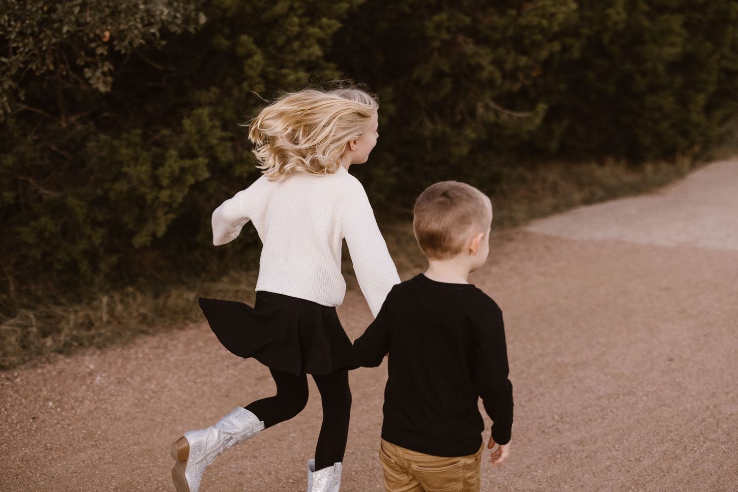 Two young siblings run hand in hand down a tree-lined dirt path together.