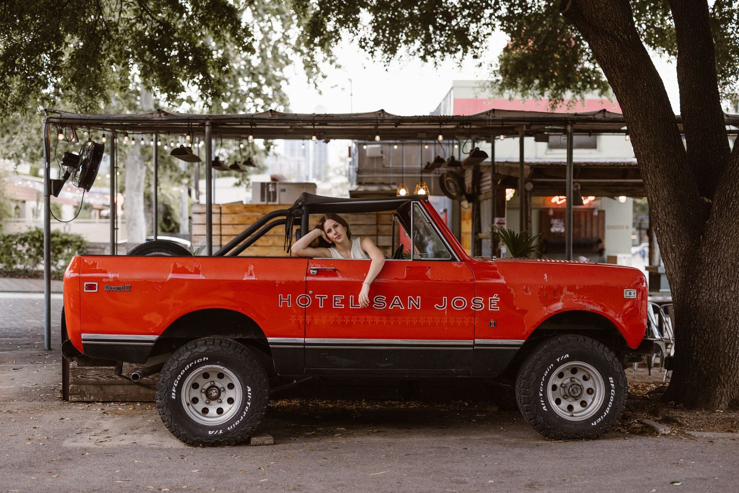 Vintage red open-top SUV with custom lettering parked under a large tree in an urban setting.