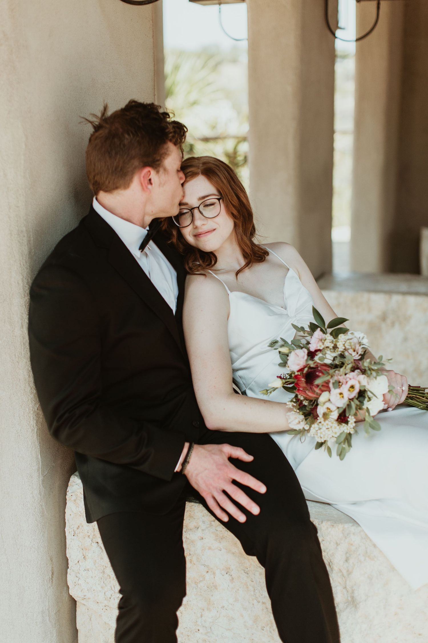 A bride and groom share a tender moment seated together, she holds a pink and white bouquet while leaning into him.