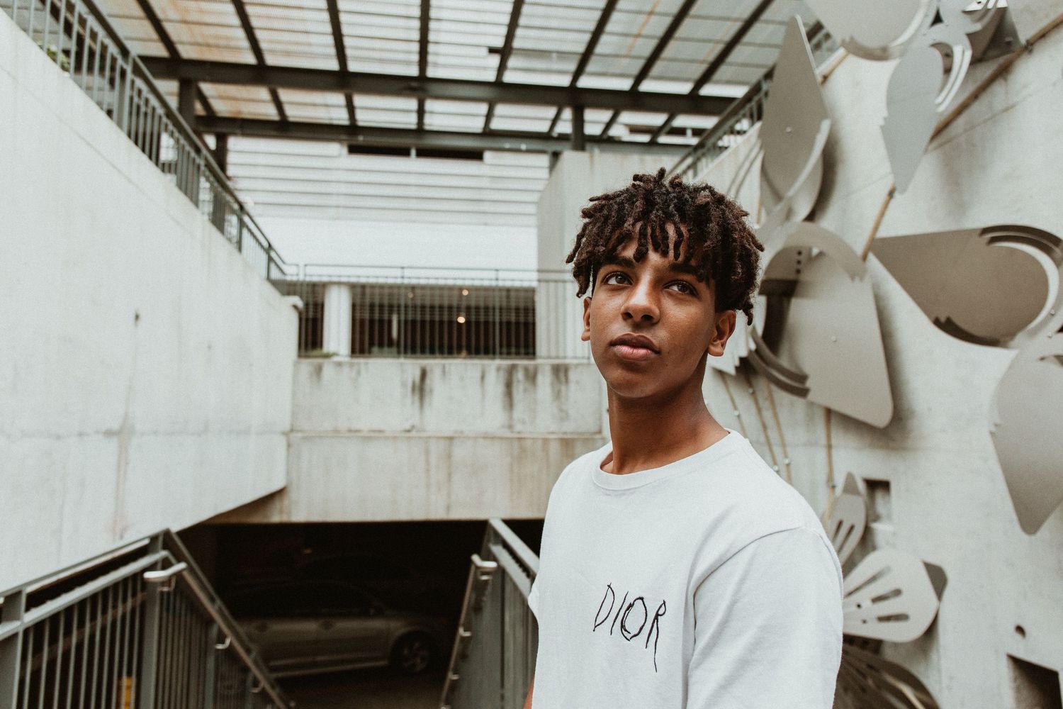 Young man in white Dior sweatshirt with short dreadlocks posing in urban concrete setting with stairs.