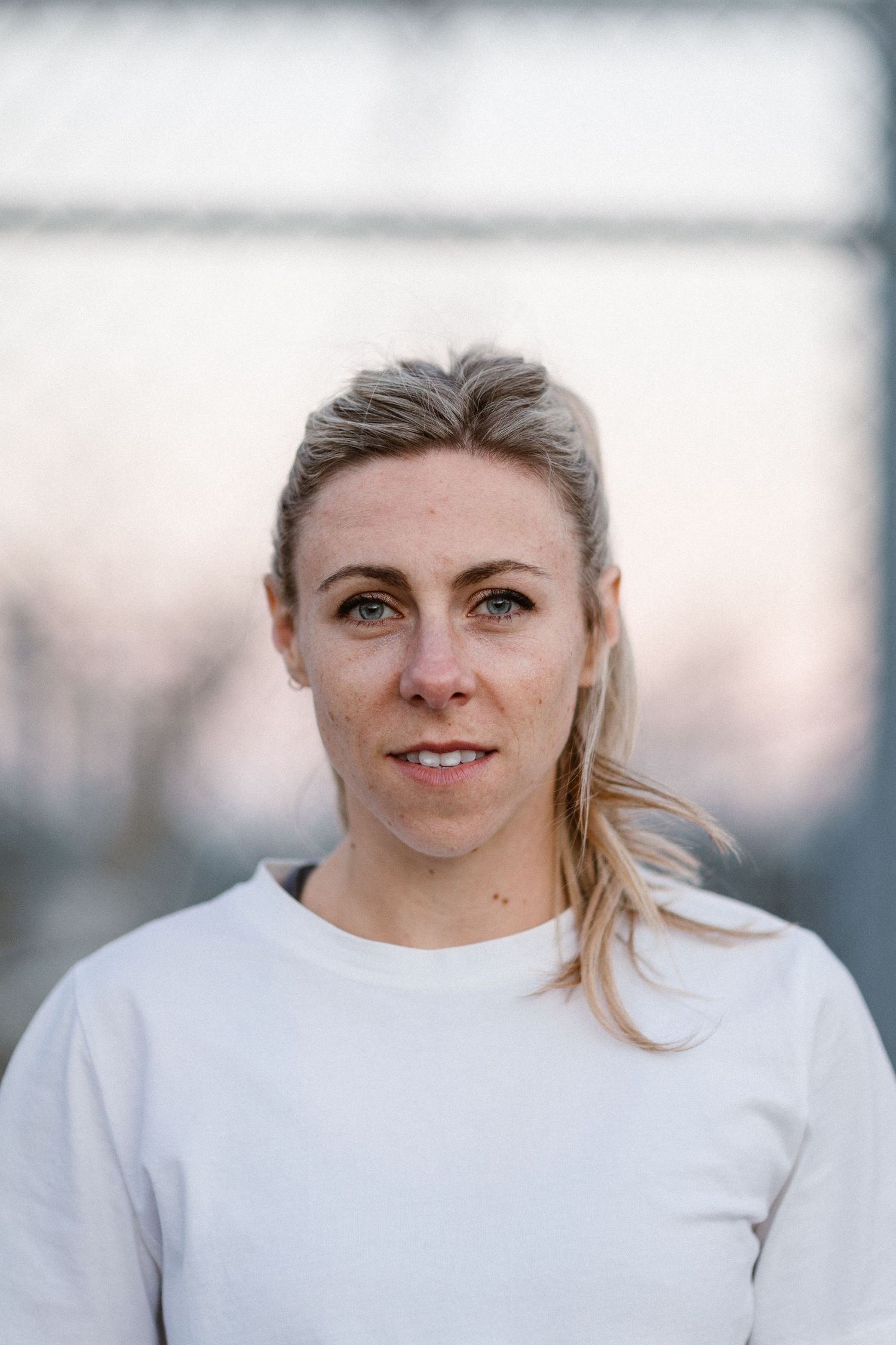 Portrait of a blonde woman in a white sweatshirt outdoors, with a blurred chain-link fence background.