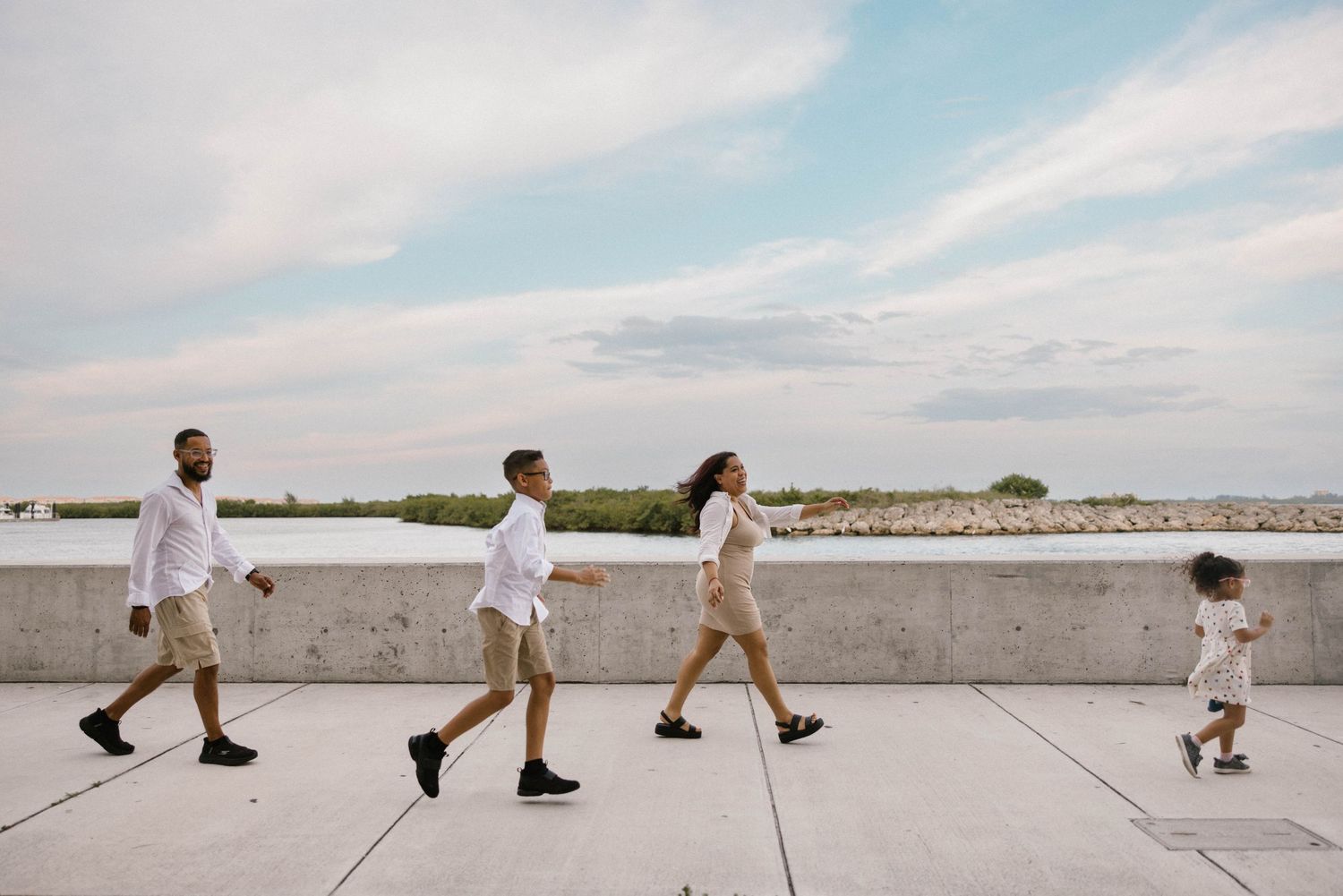 Family of four walking in a line along a waterfront promenade under a cloudy sky in matching white outfits.