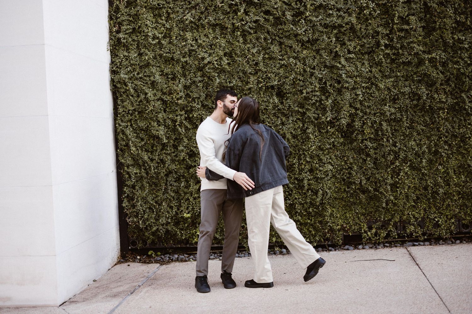 Romantic couple sharing a kiss against a white wall covered with lush green ivy outdoors.