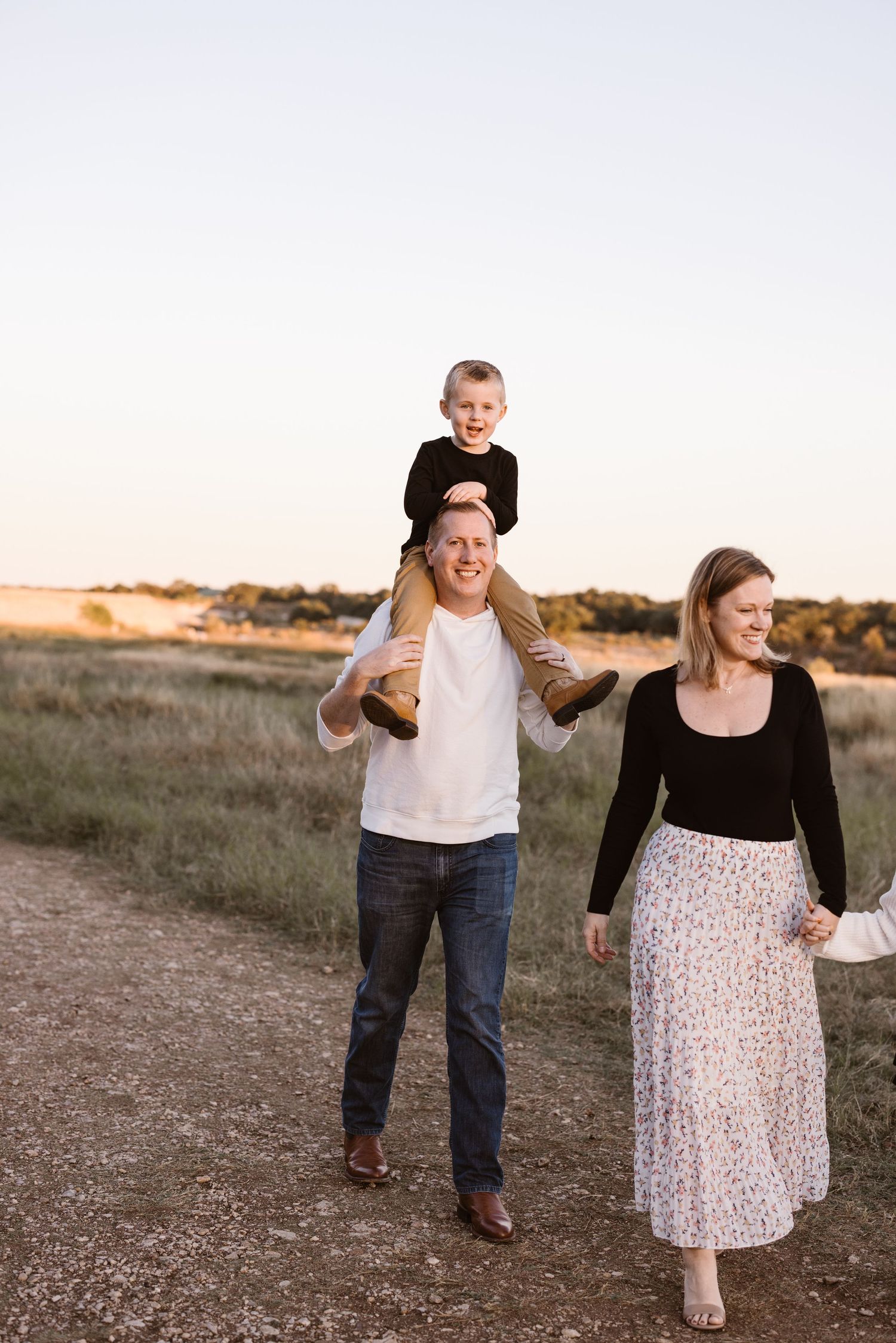 A happy family walks along a dirt path at sunset, with a young boy sitting on his father's shoulders.