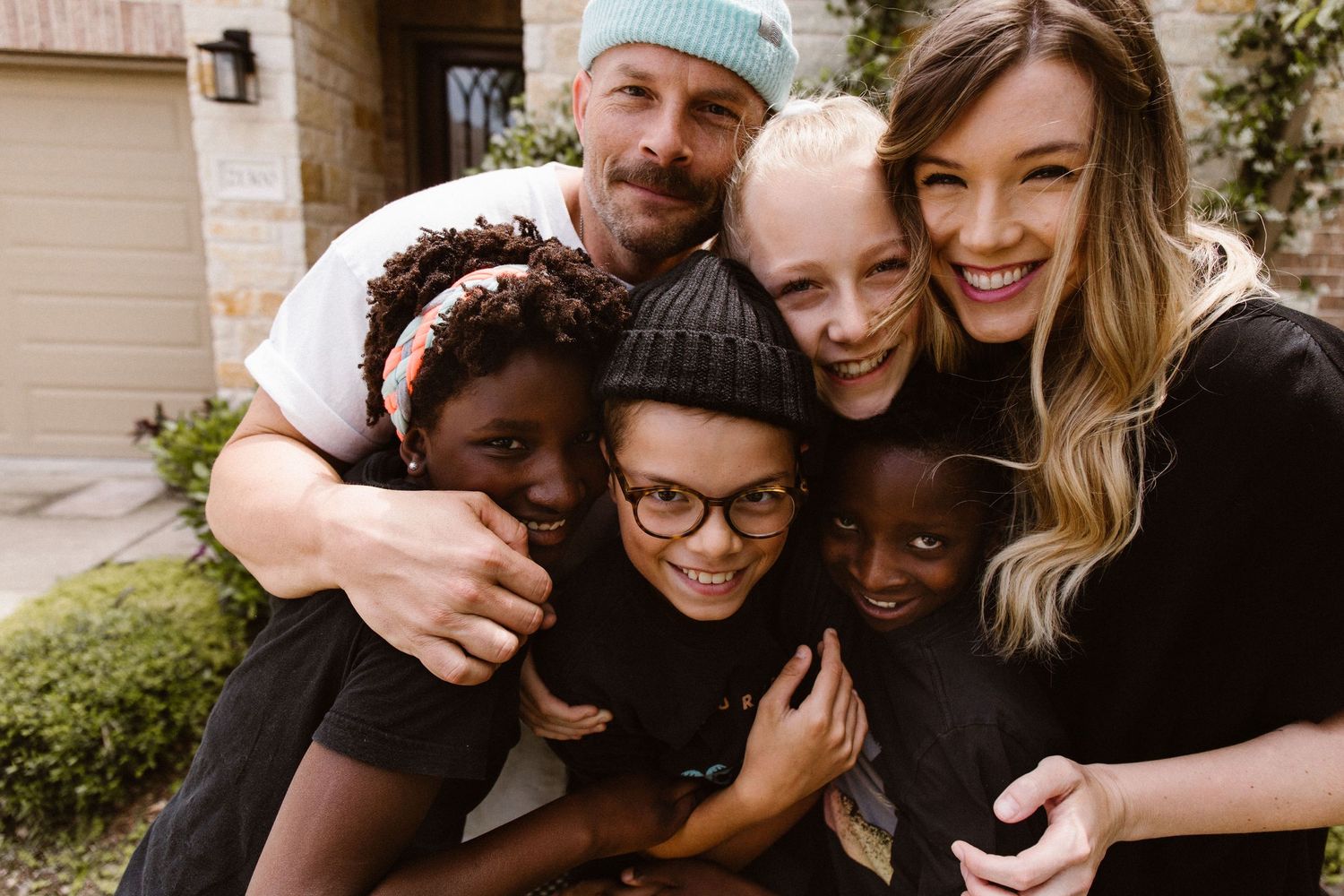 A diverse family of six smiling and hugging together outside their home.