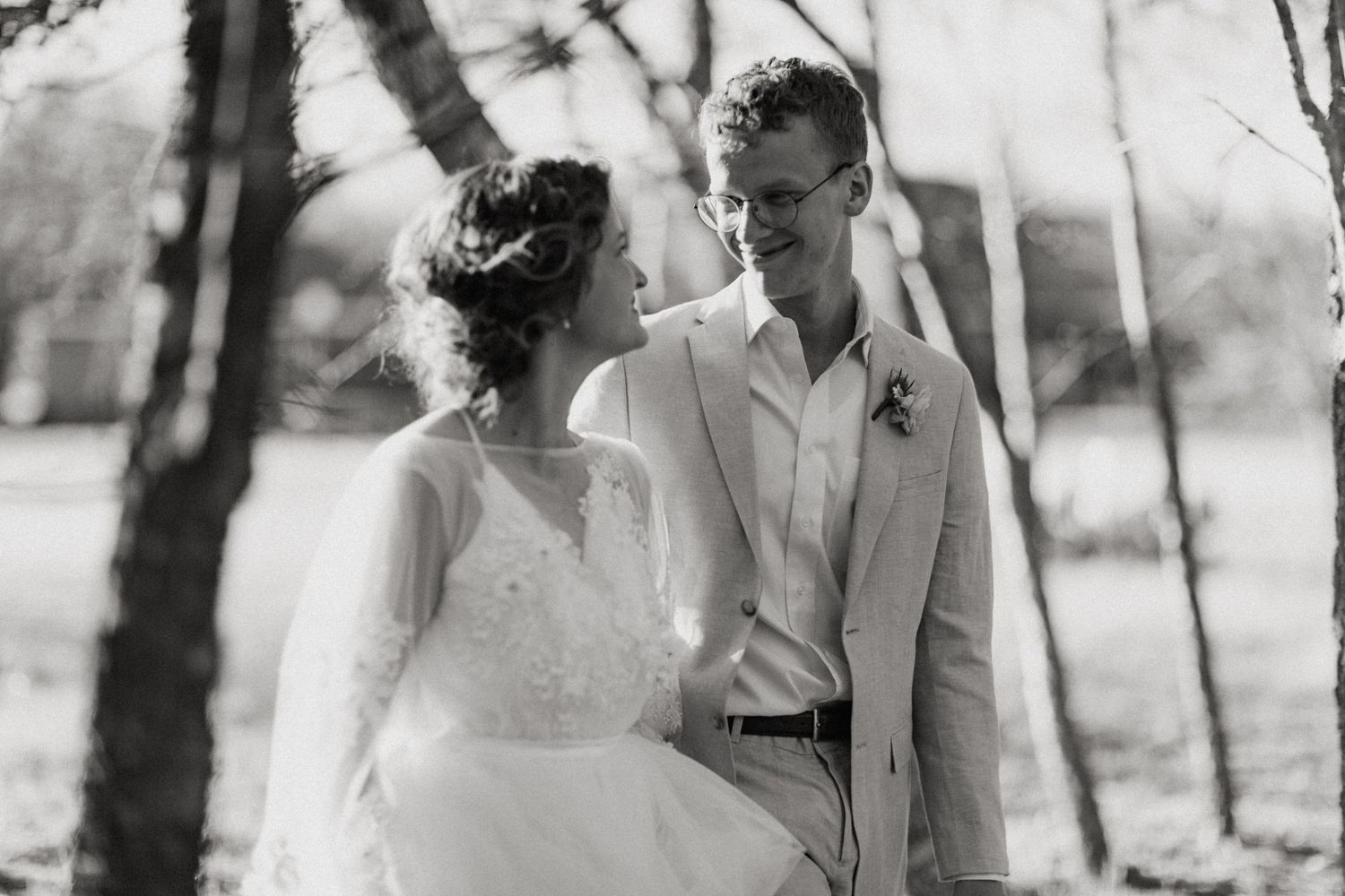 Black and white photo of smiling bride and groom walking together among trees on wedding day.