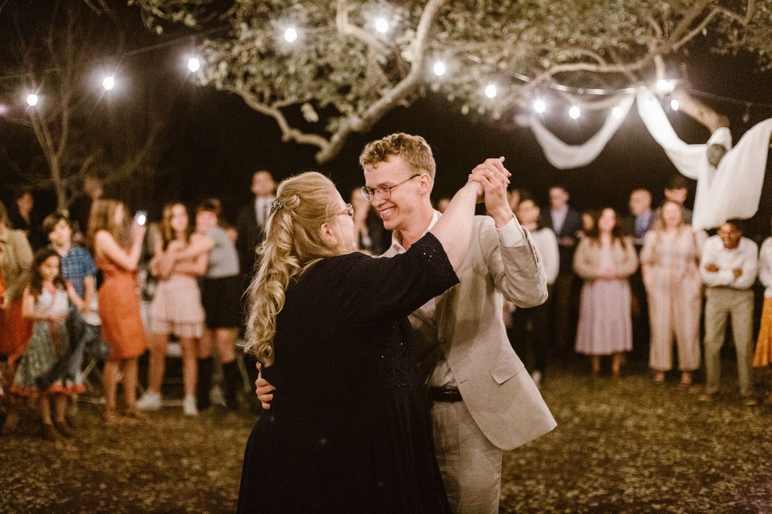 Groom dancing with mother at outdoor wedding reception surrounded by guests under string lights.