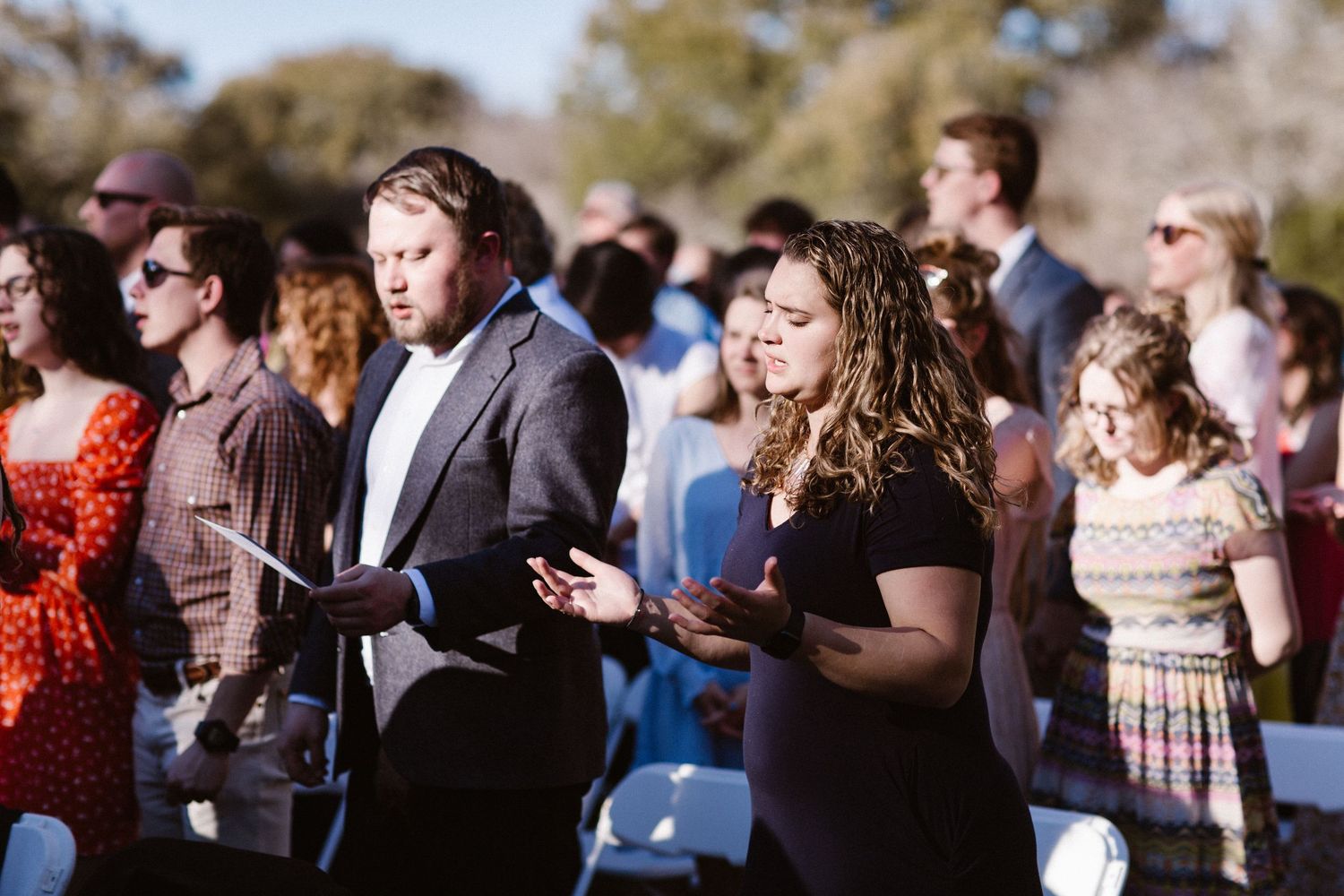 Wedding guests holding programs and singing during an outdoor ceremony on a sunny day.