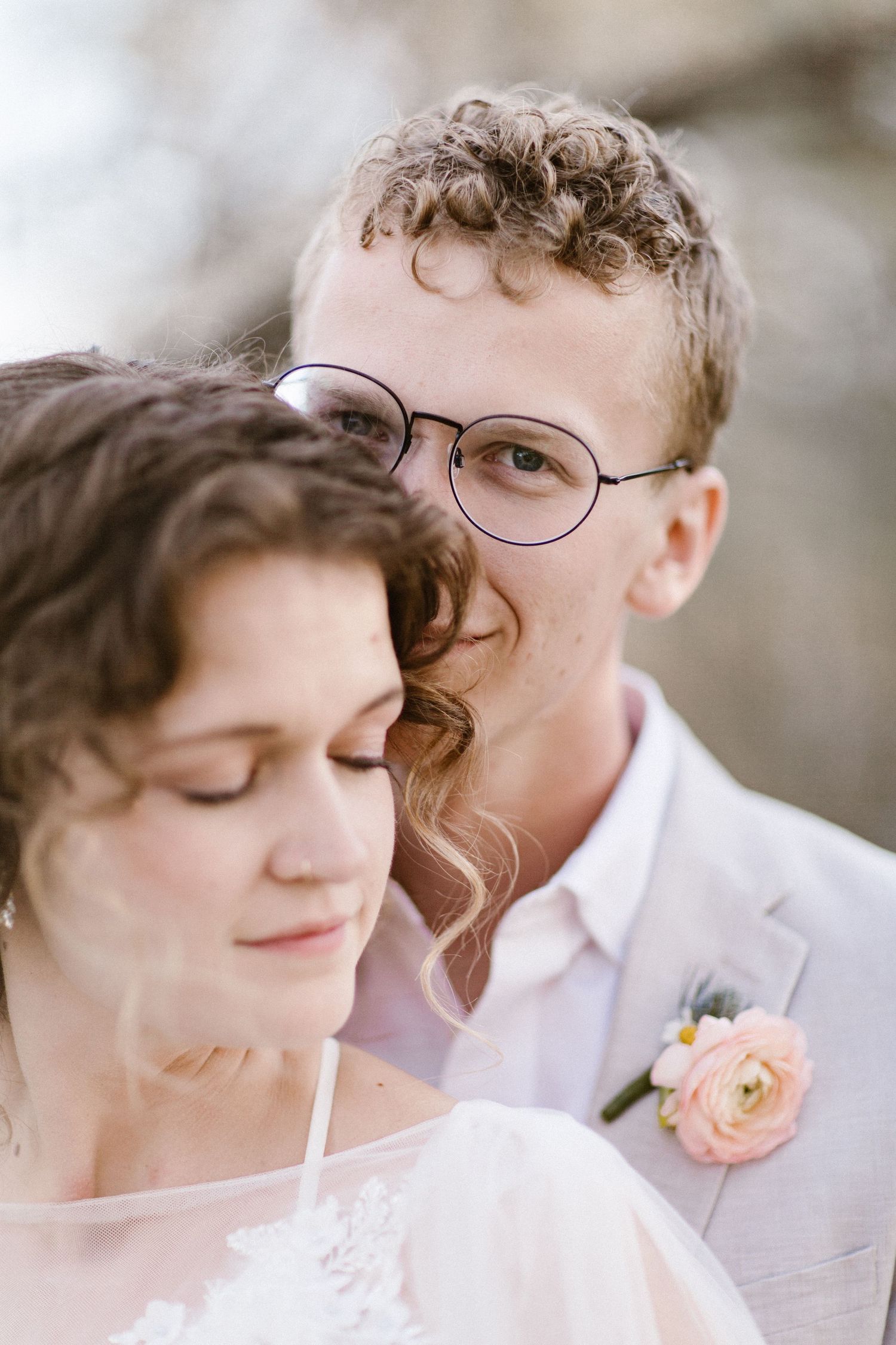 Close-up portrait of groom with glasses and curly hair embracing bride in white lace wedding dress.