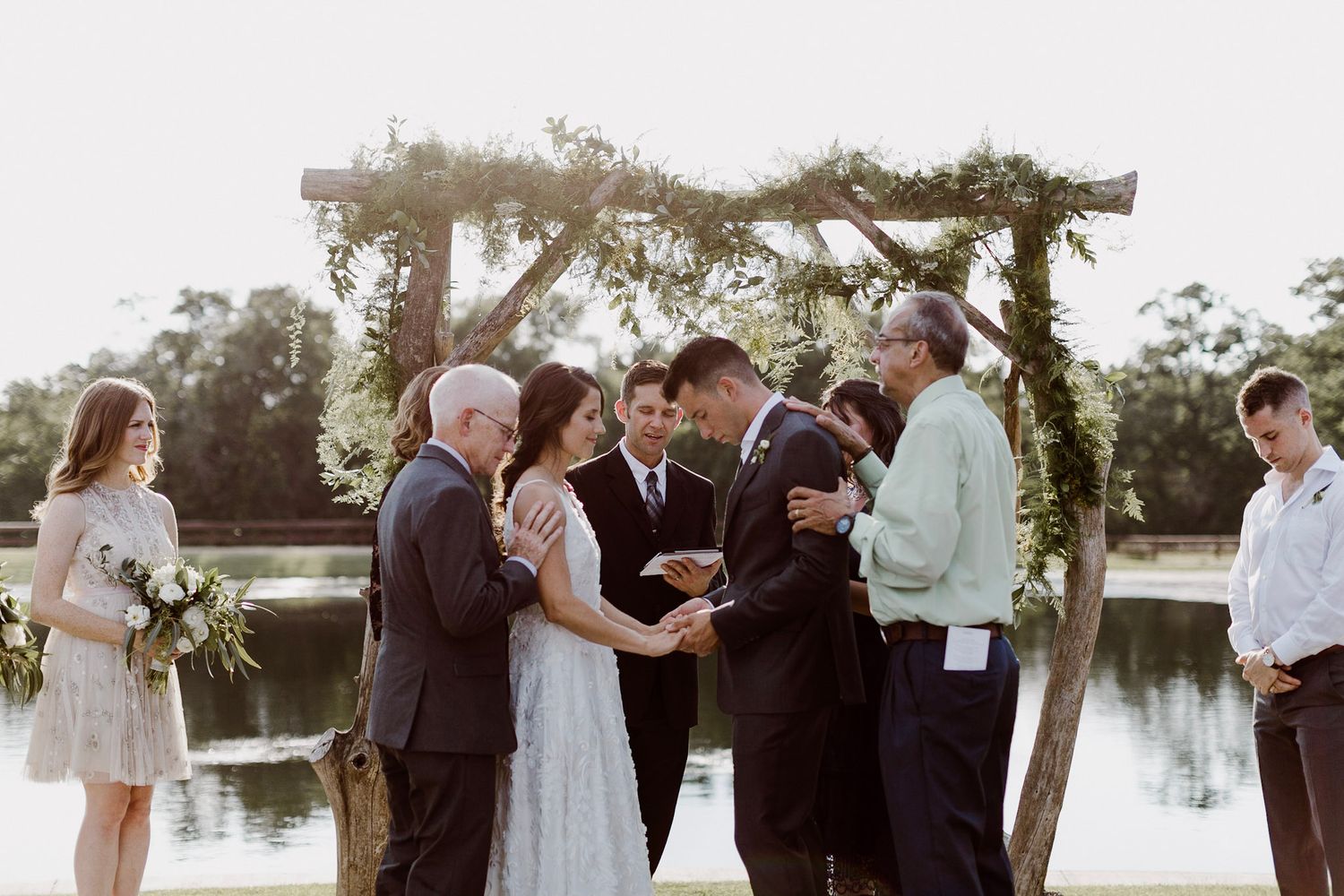Intimate outdoor wedding ceremony at waterside rustic wooden arch with small wedding party and officiant.