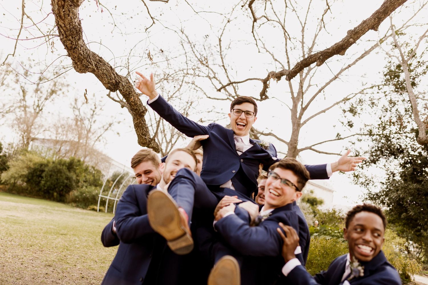 Groomsmen in navy suits lifting and carrying groom outdoors in a joyful wedding celebration photo.