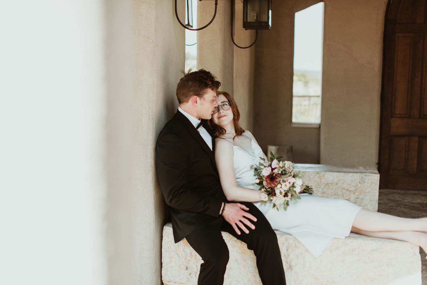 A bride and groom sit together on a stone bench, sharing a tender moment, with the bride holding a floral bouquet.