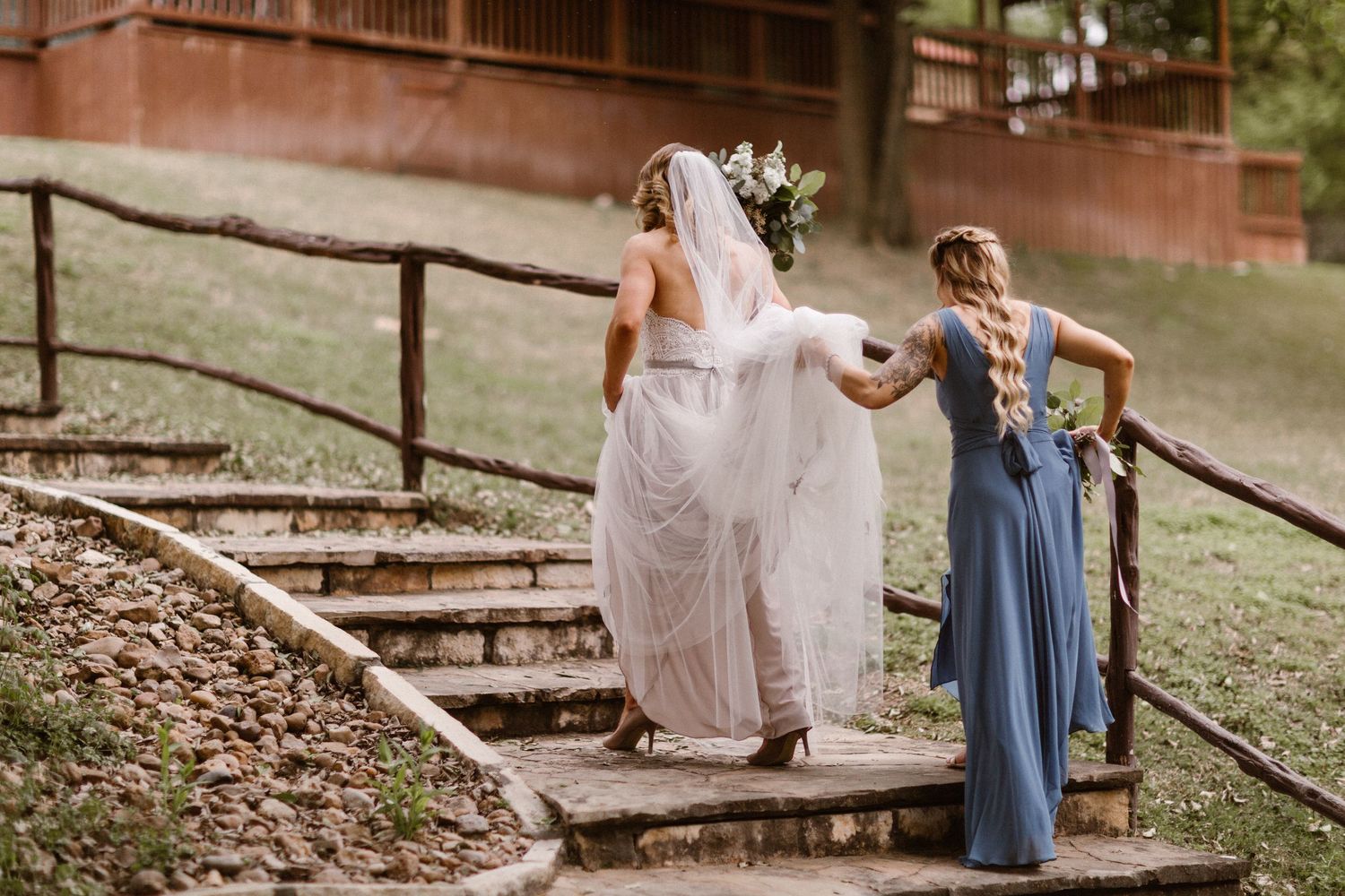 A bridesmaid in blue helps a bride in a white gown walk up outdoor stone steps.