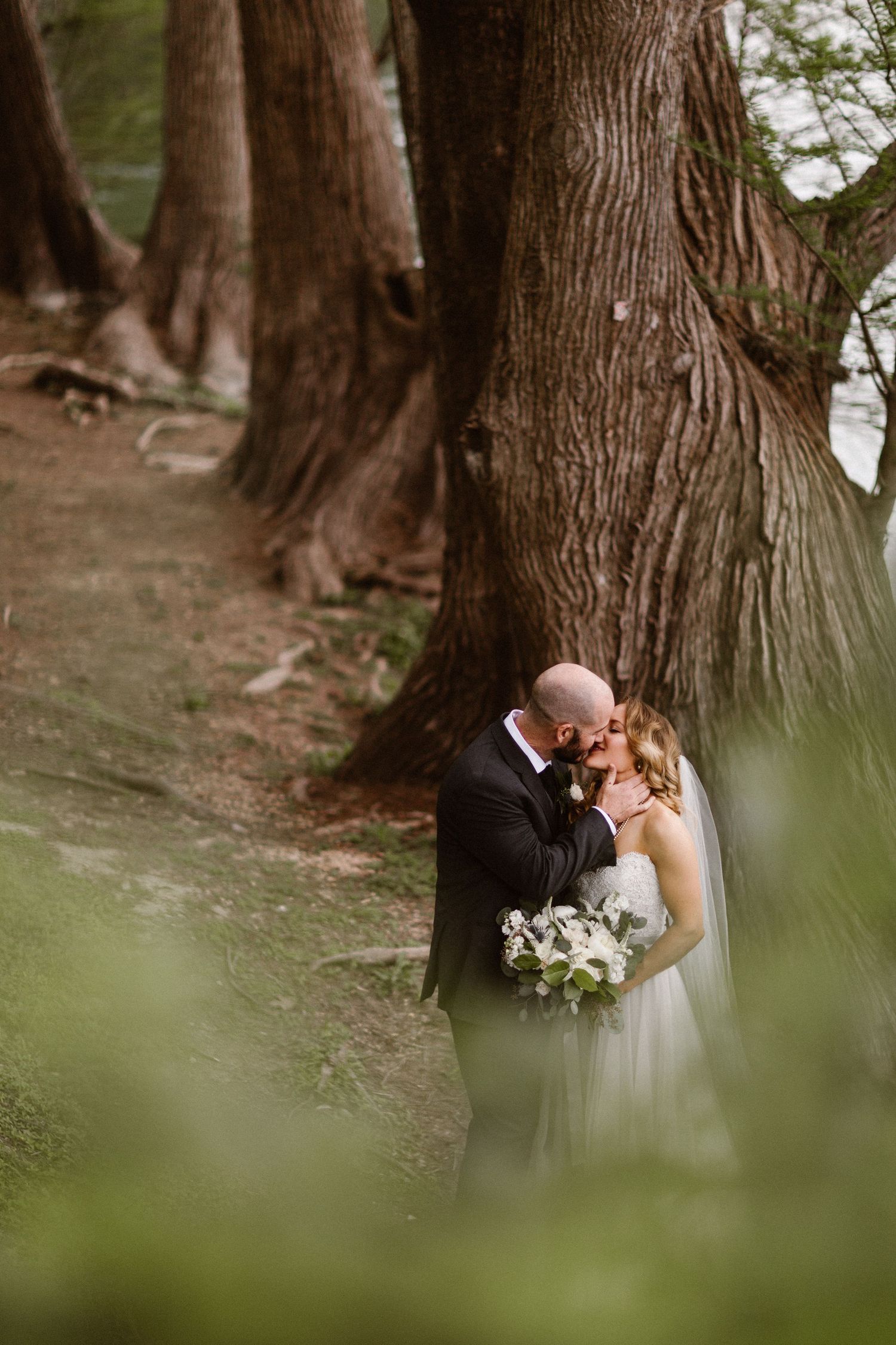 A bride and groom share a kiss among tall trees in a lush forest setting.