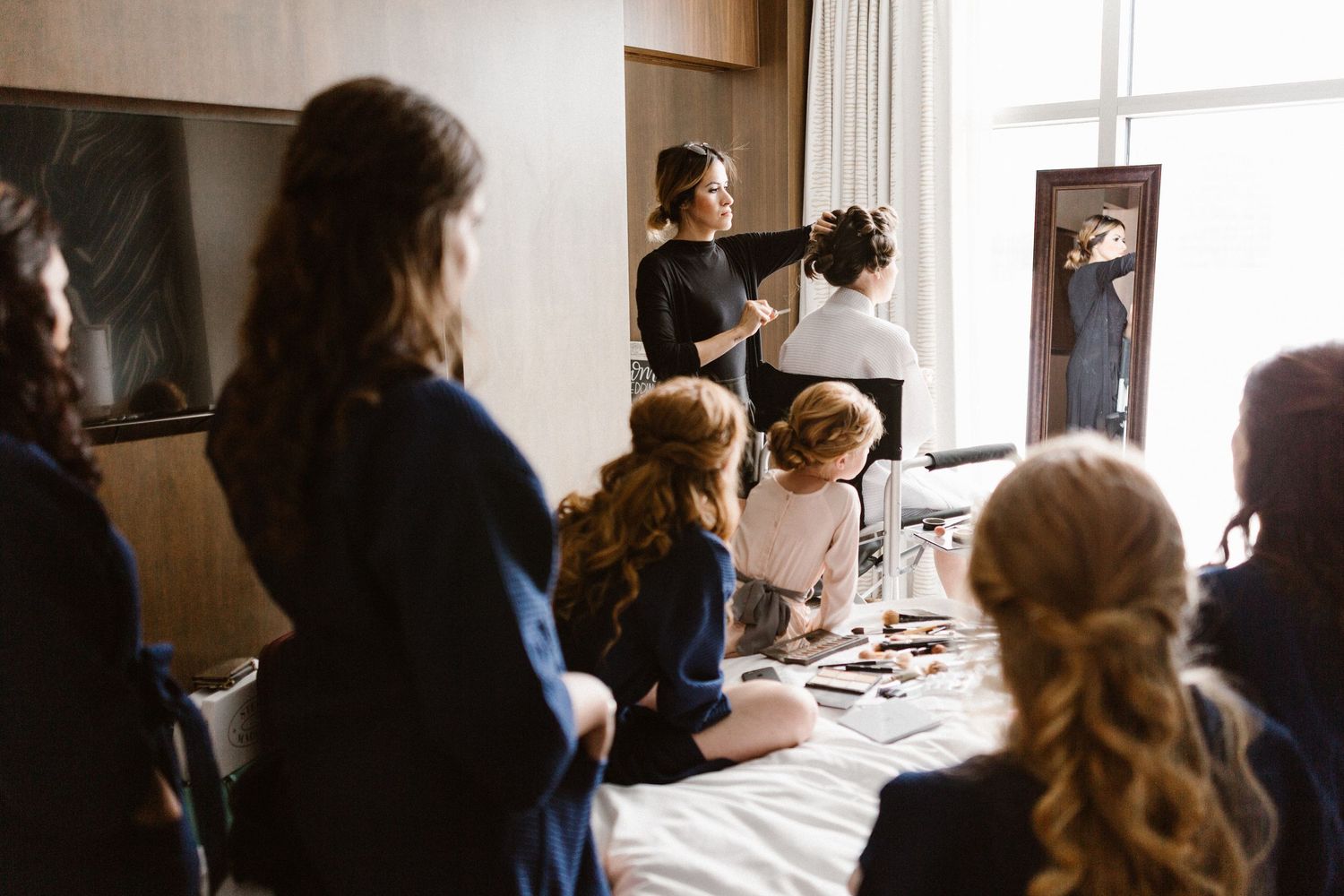 Bridesmaids in robes watching hairstylist prepare bride in bright hotel room before wedding.