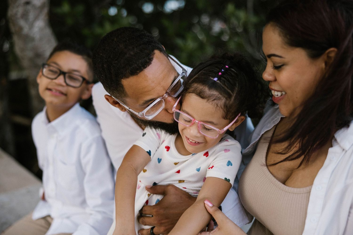 Close-up of a joyful family with father kissing young daughter wearing pink glasses while mother and son smile nearby.