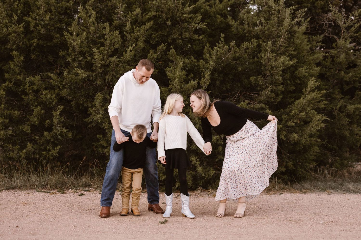 Playful family of four laughing together on a dirt path with evergreen trees behind.