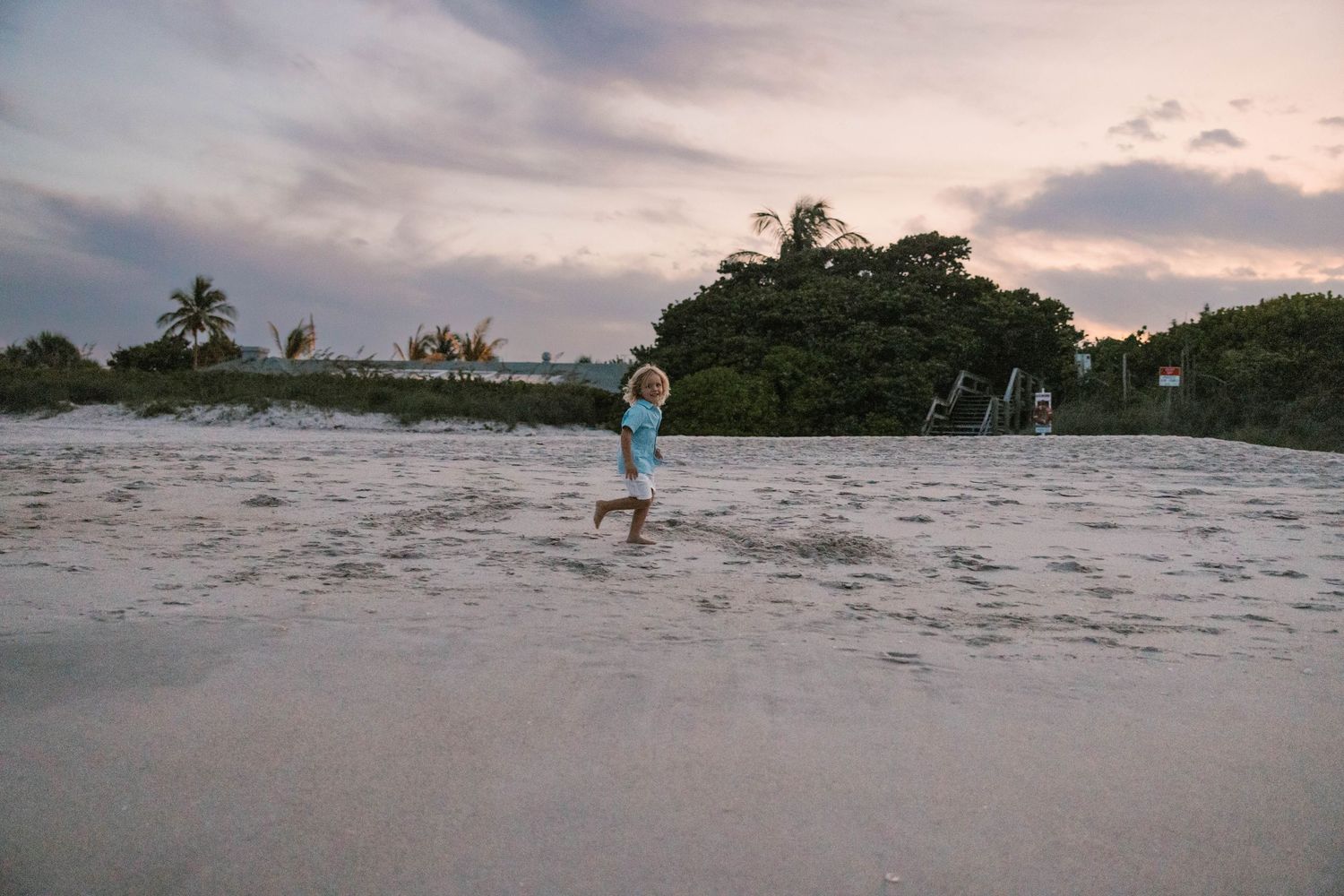 Young child running on a sandy beach at sunset with trees in background.