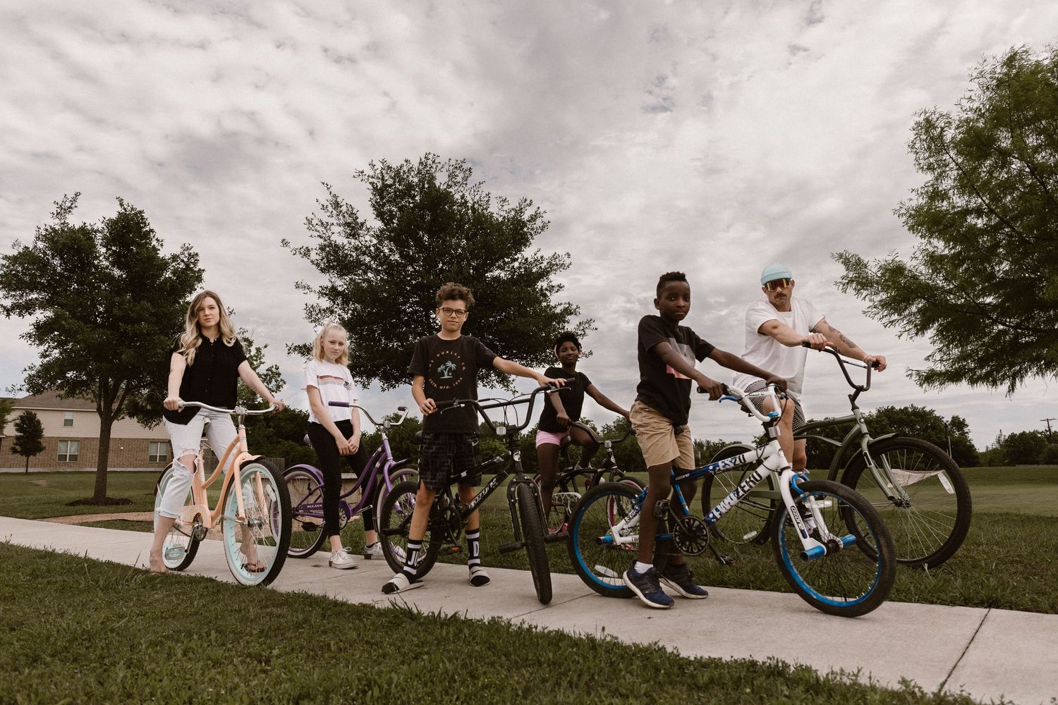 Group of six young people posing with bicycles on a suburban sidewalk.